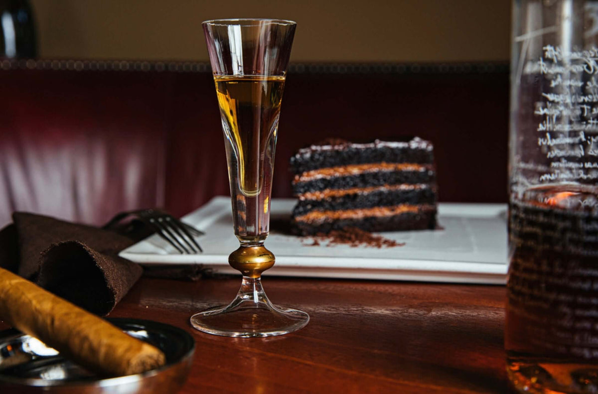 A glass of Tears of Llorona Extra Añejo Tequila beside a chocolate cake and cigar on a wooden table.
