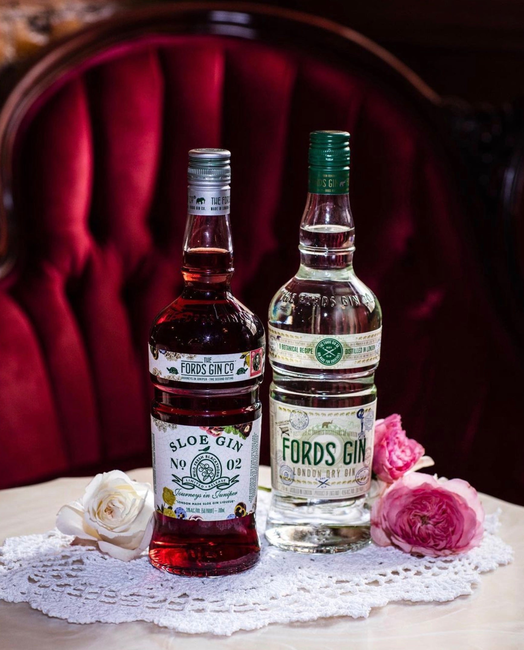 Fords Sloe Gin and London Dry Gin bottles on lace doily with flowers, set against a vintage red background.