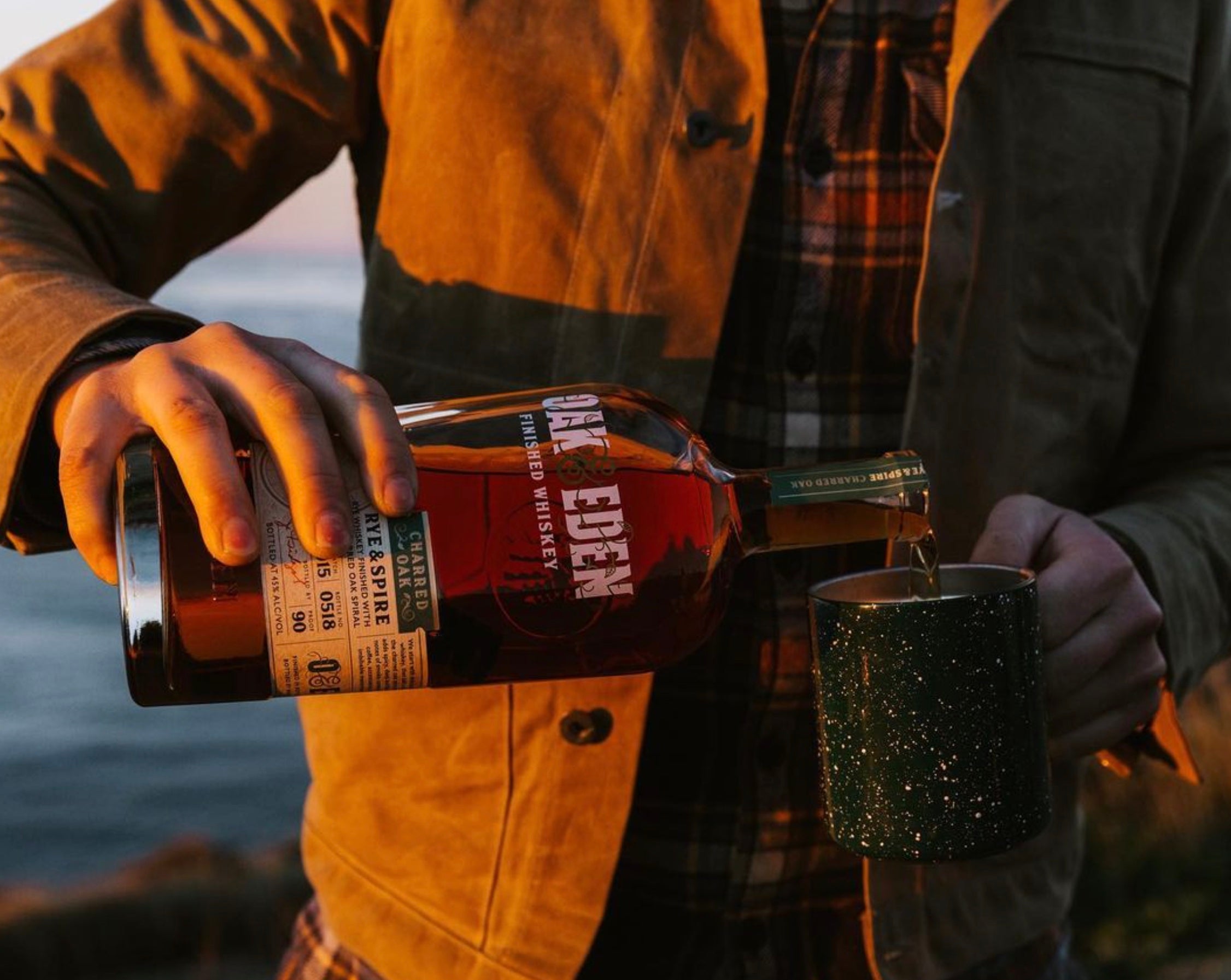 Pouring Oak & Eden Rye & Spire Whiskey into a green mug by the water at dusk.