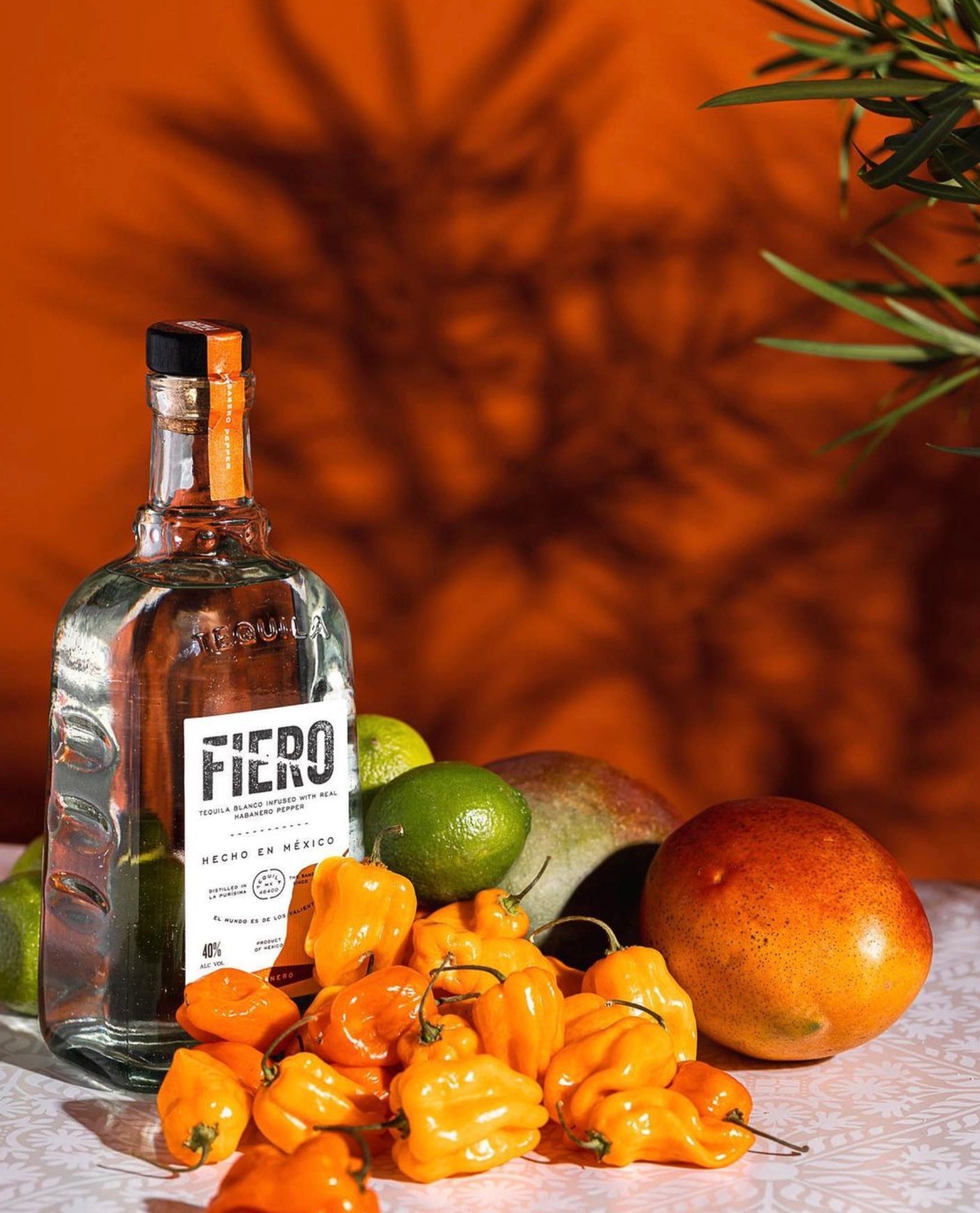 Fiero Habanero Tequila bottle surrounded by habanero peppers, limes, and mangoes on a vibrant orange backdrop.