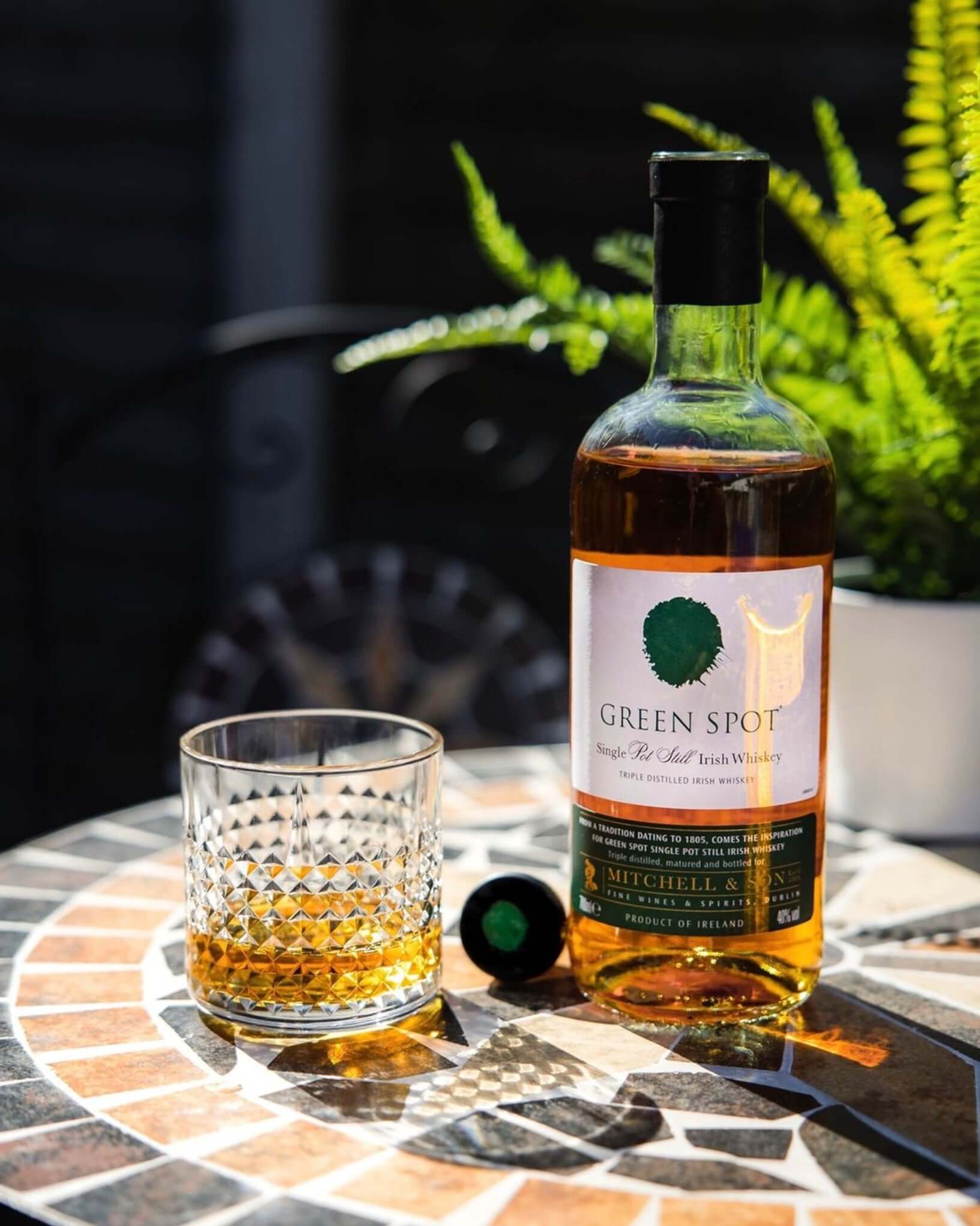Green Spot Irish Whiskey bottle beside a glass of whiskey on a patterned table with greenery in the background.