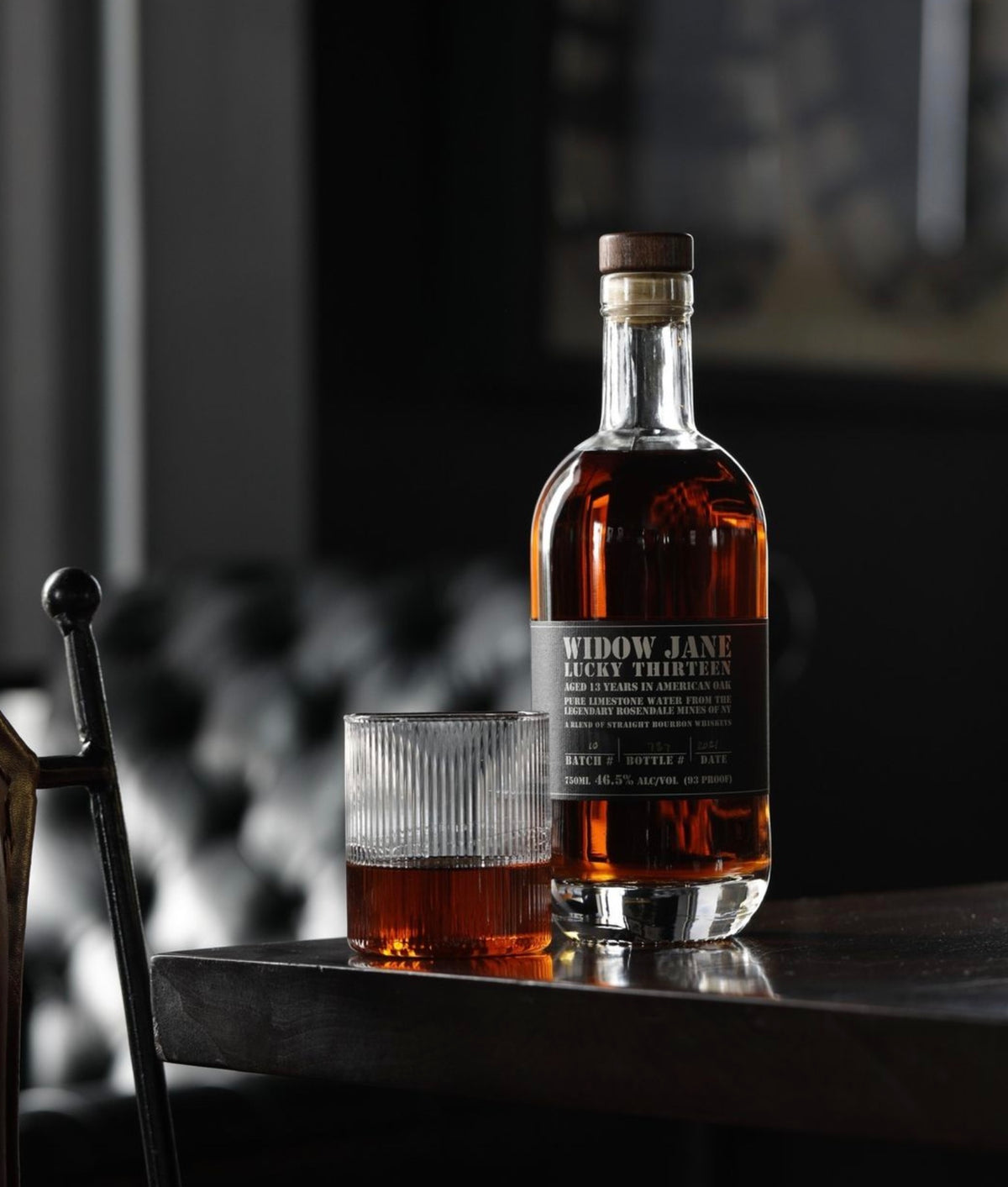 Widow Jane Lucky Thirteen Bourbon bottle with glass on a table, highlighting rich amber color and craftsmanship.