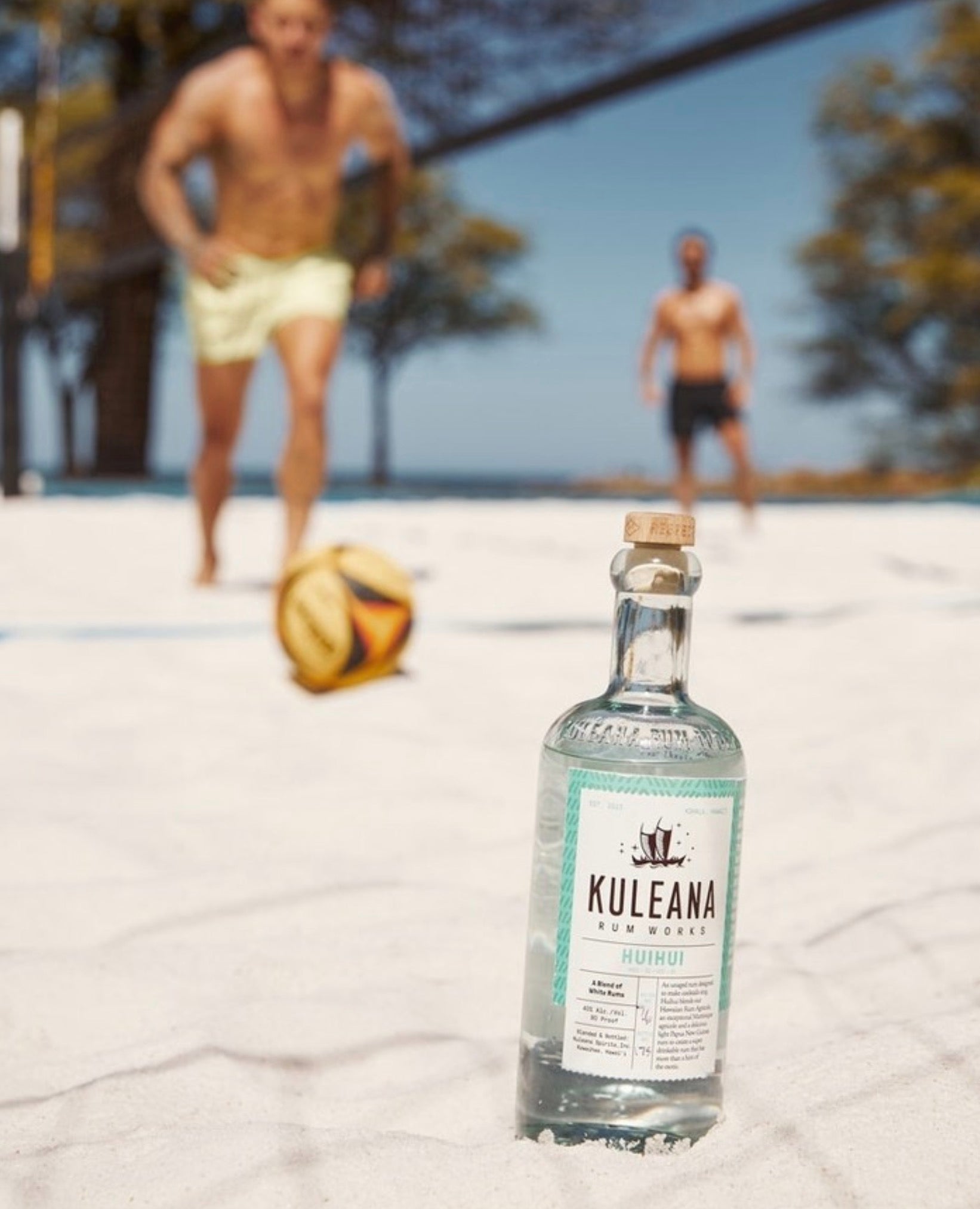 Kuleana Huihui Rum bottle on beach with volleyball players in the background, highlighting tropical vibe and enjoyment.