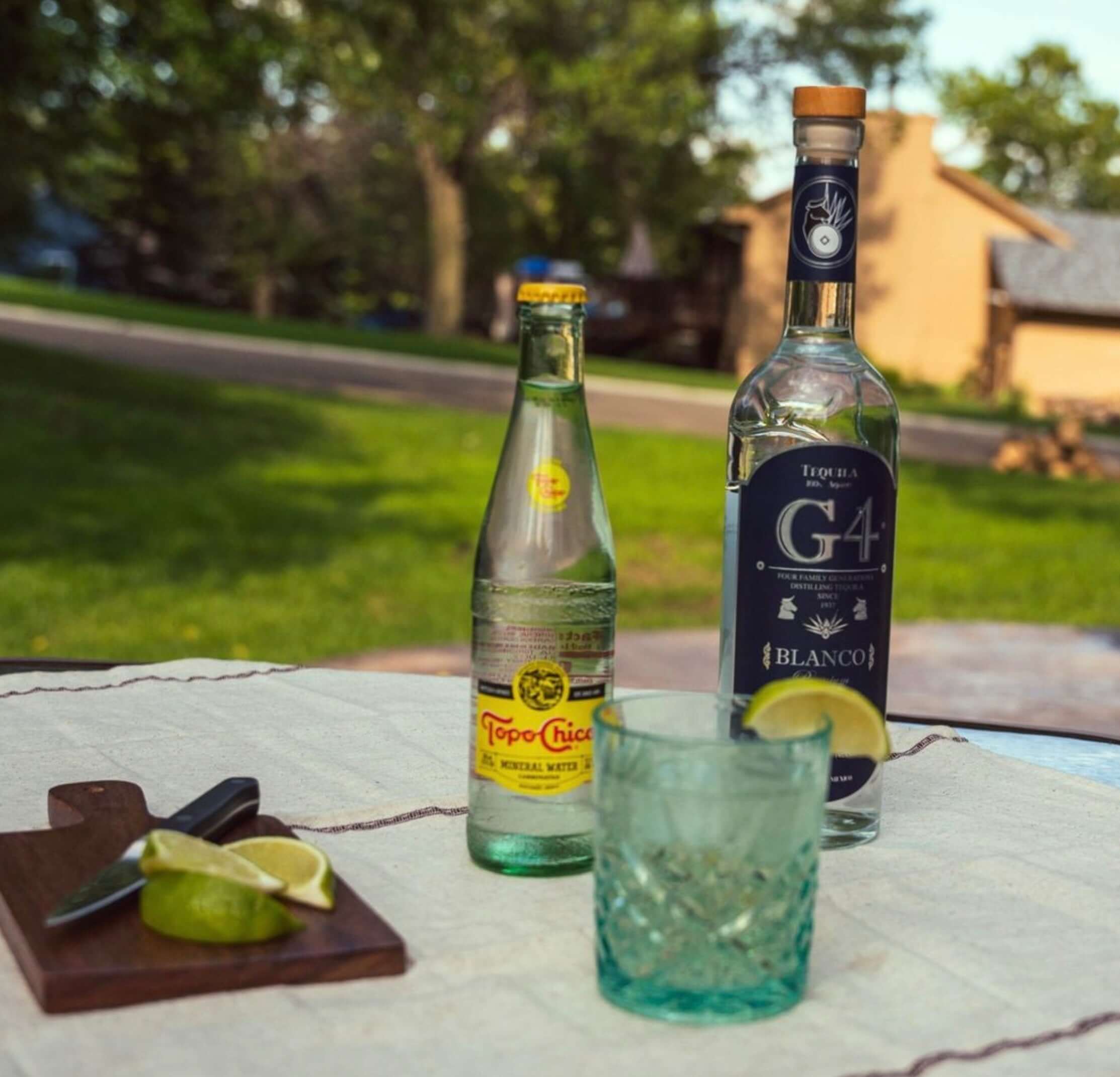 G4 Blanco Tequila bottle with lime, cocktail glass, and Topo Chico on an outdoor table.