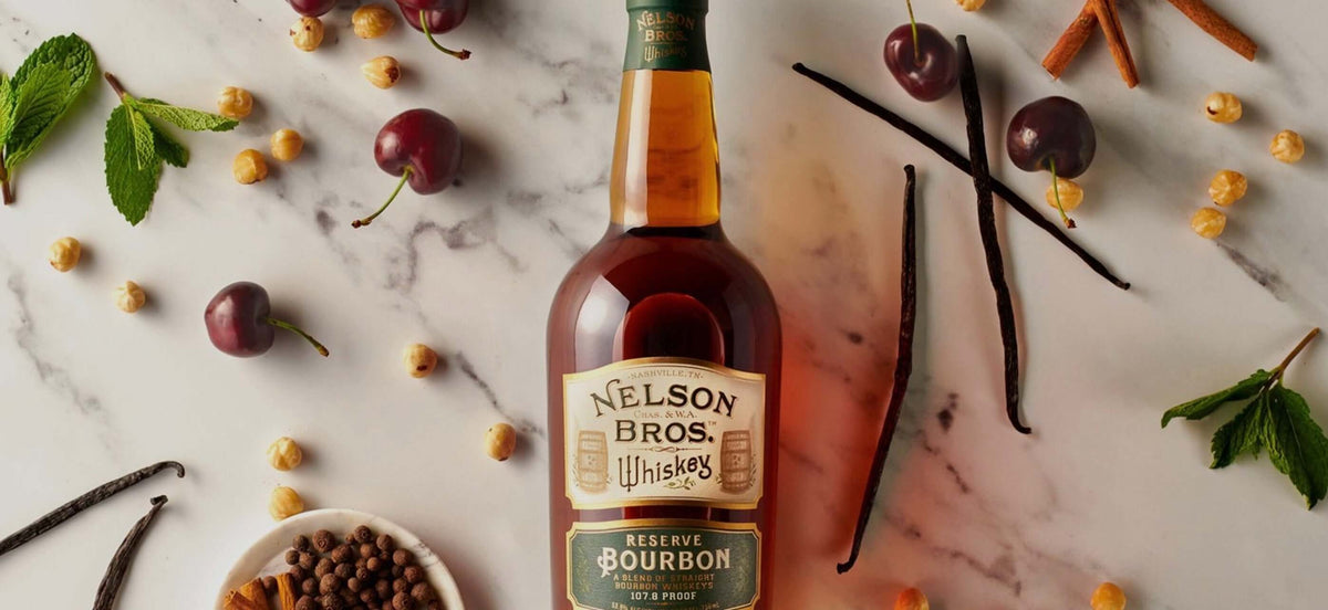 Nelson Brothers Reserve Bourbon bottle surrounded by cherries, mint, vanilla, and spices on a marble surface.