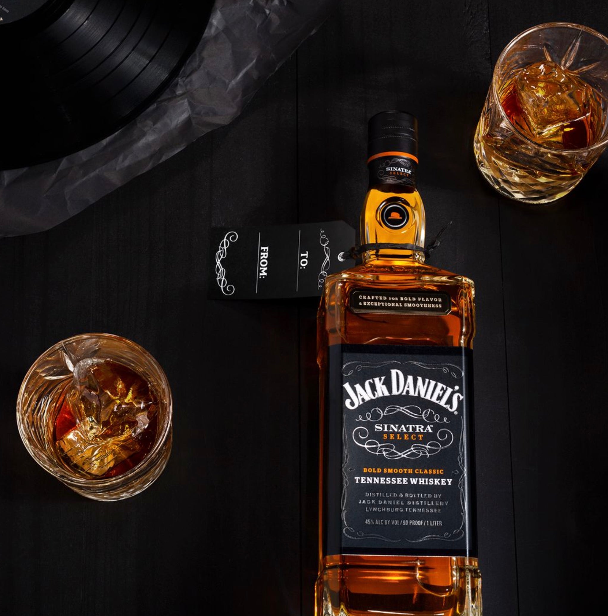 Jack Daniel's Sinatra Select whiskey bottle with glasses of whiskey and a vinyl record on a dark wooden table.