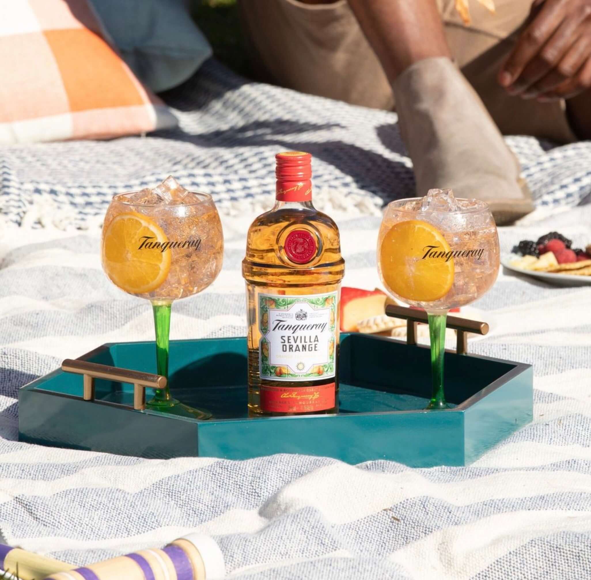 Tanqueray Sevilla Orange Gin with two cocktails on a picnic blanket, garnished with fresh orange slices.