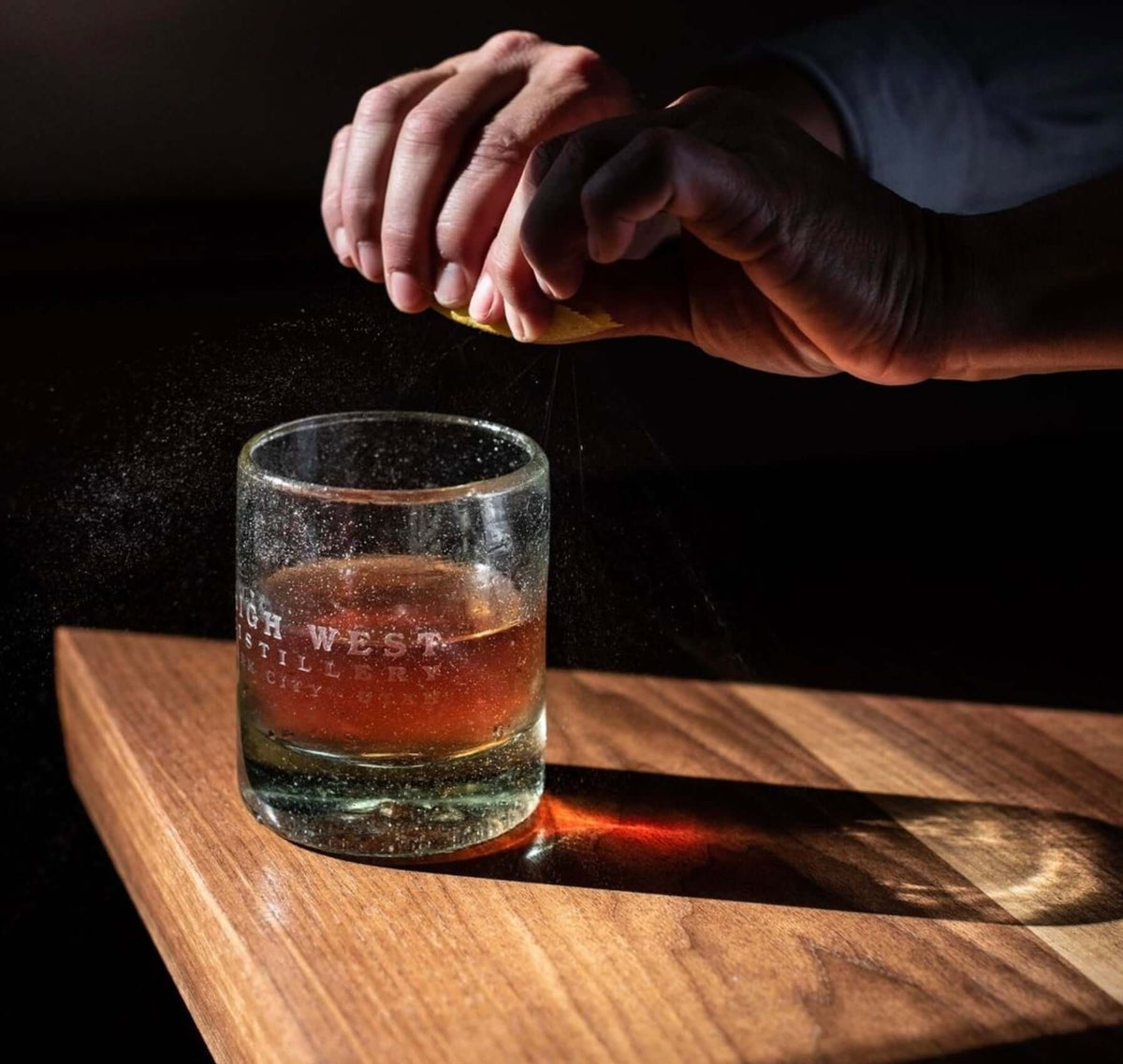Person squeezing lemon over a glass of High West Bourbon on a wooden surface, creating an aromatic cocktail.
