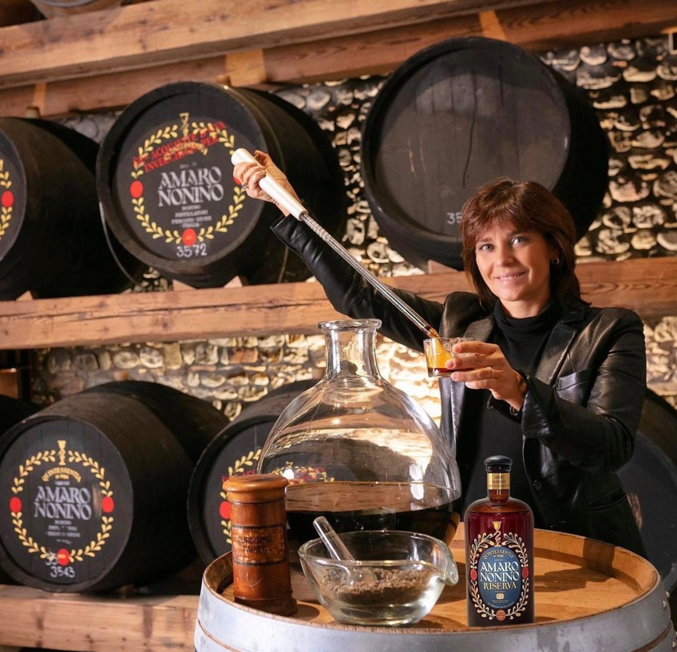 Woman serving Amaro Nonino Quintessentia Riserva in a distillery with wooden barrels in the background.