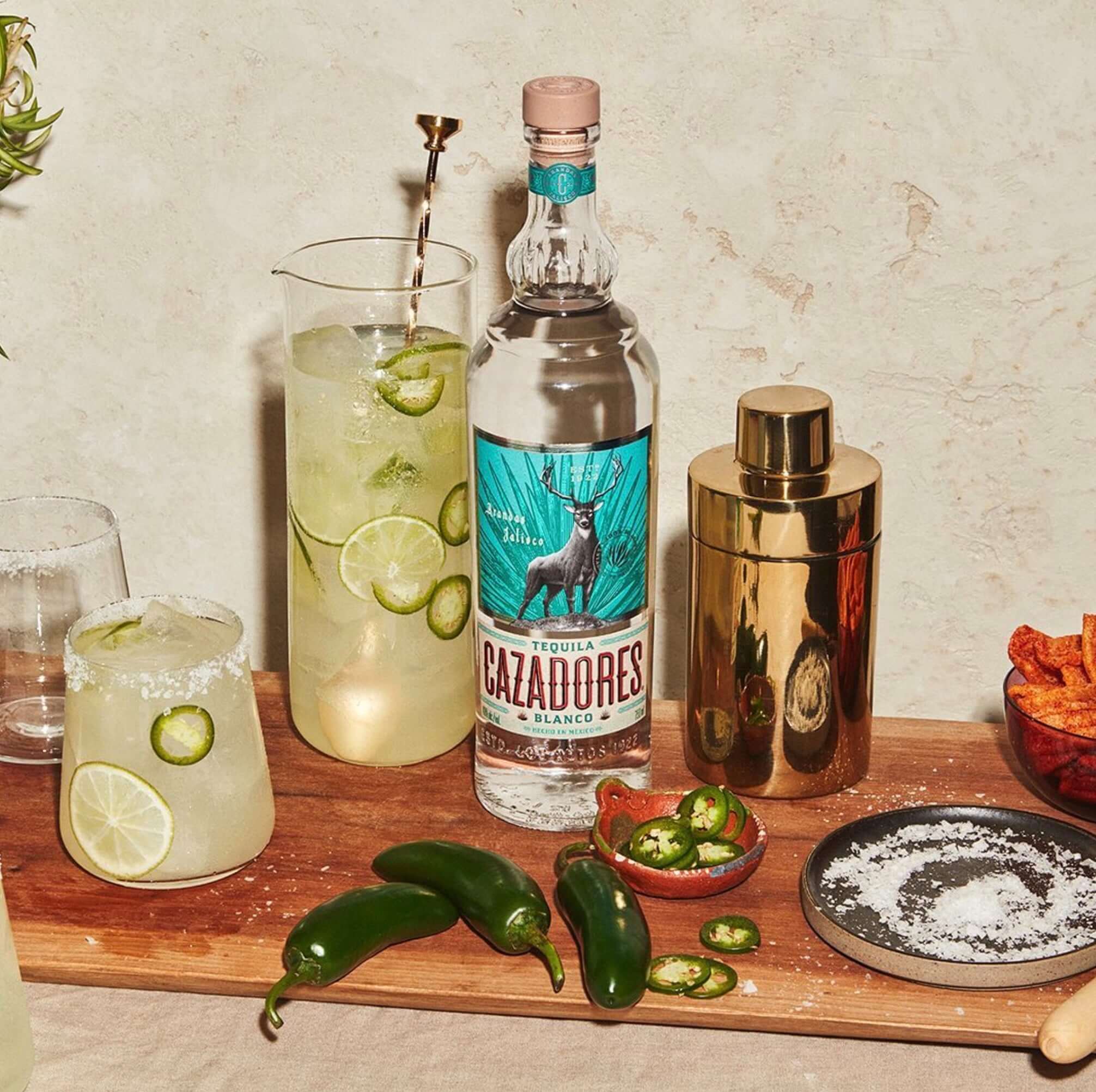 Cazadores Blanco Tequila bottle with cocktails, jalapeños, and lime on a wooden tray, perfect for gatherings.