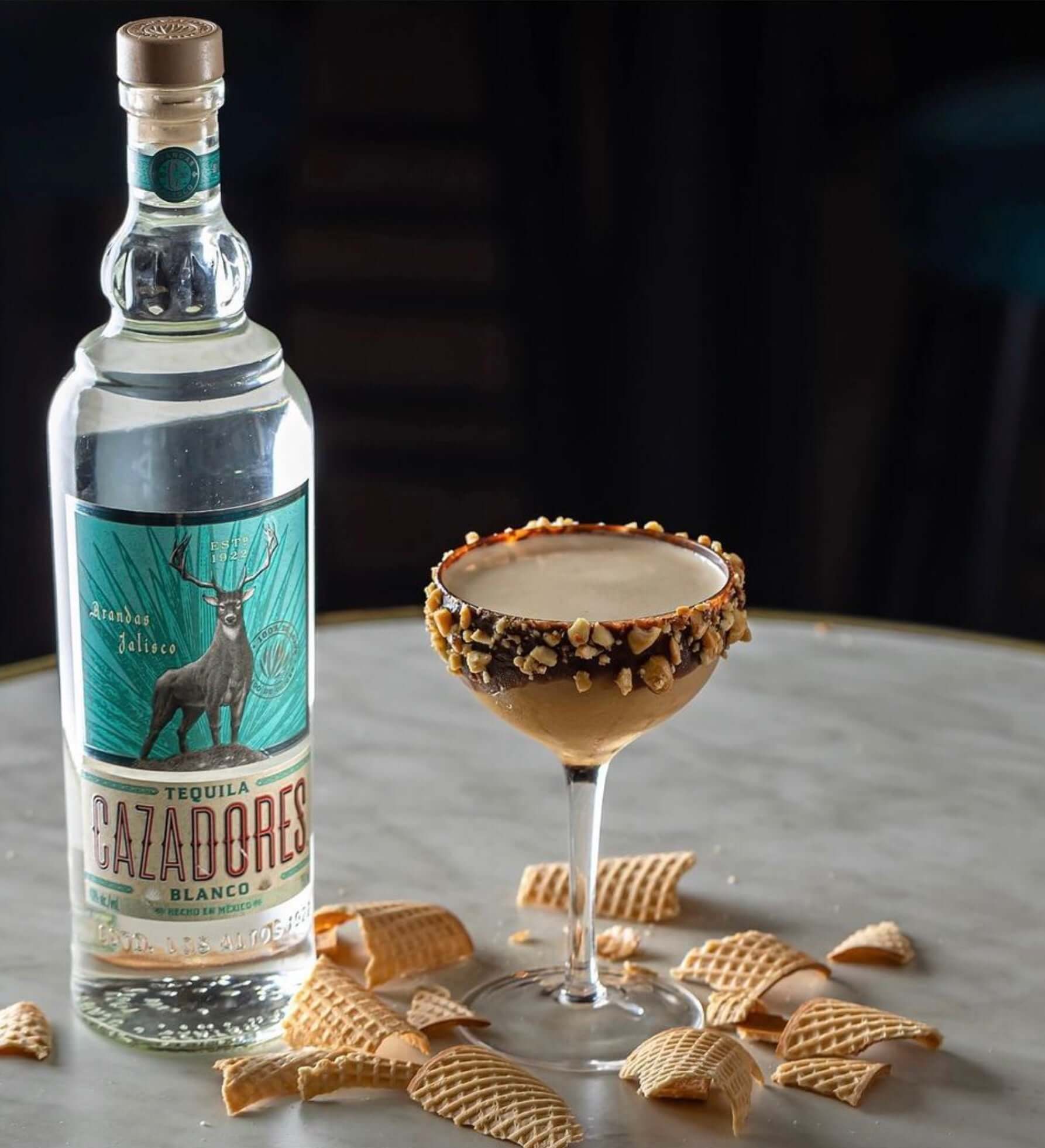 Cazadores Blanco Tequila bottle beside a cocktail garnished with nuts on a marble surface, showcasing vibrant flavors.