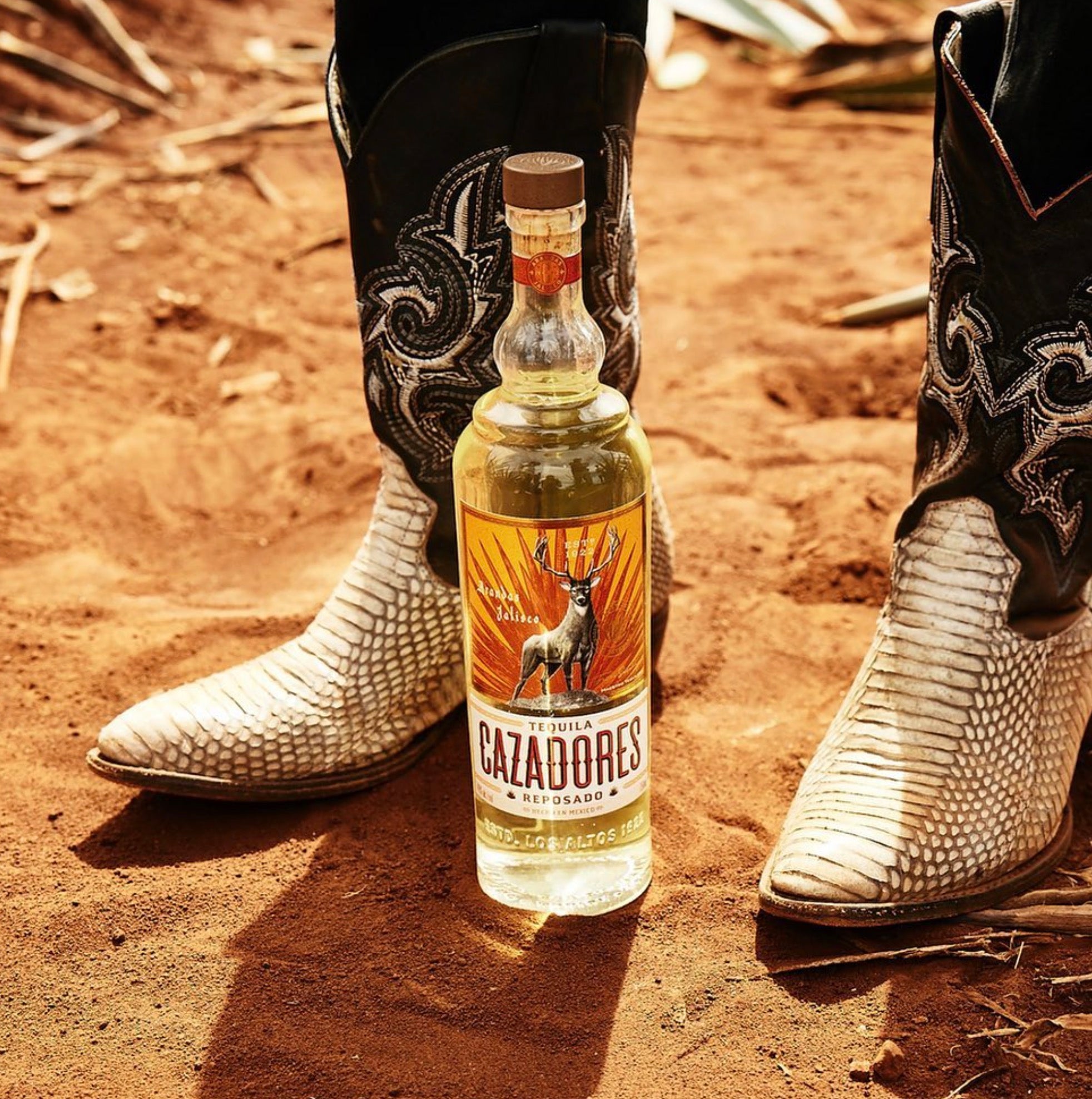 Cazadores Reposado Tequila bottle in front of cowboy boots on rustic ground, showcasing its agave and woody flavors.