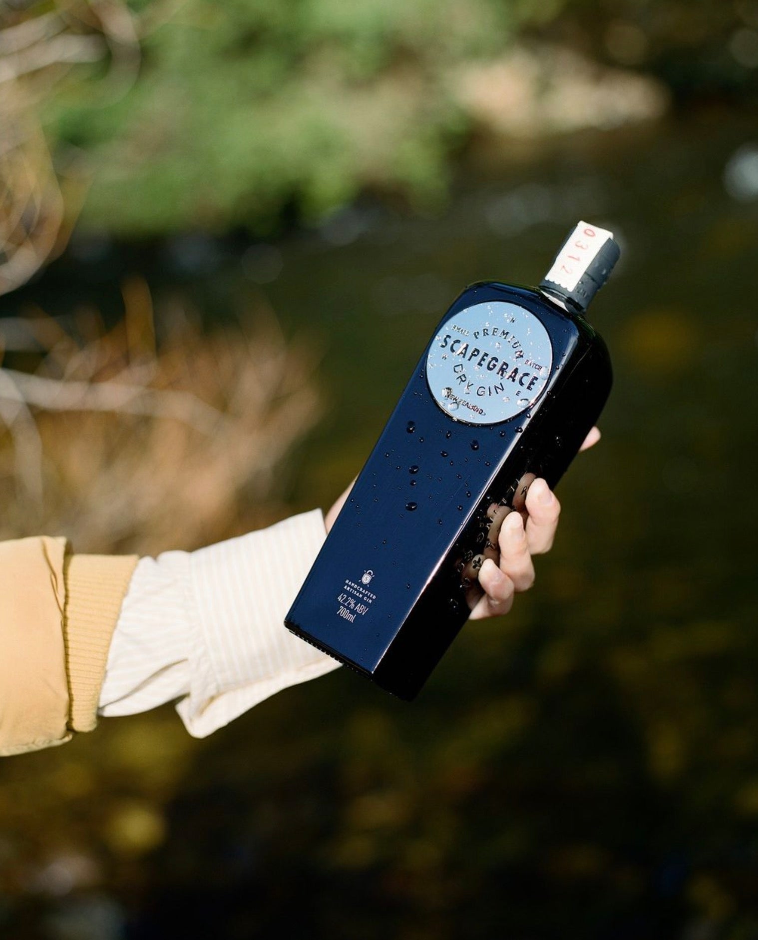 Bottle of Scapegrace Dry Gin from New Zealand held near a river, showcasing its elegant design and branding.