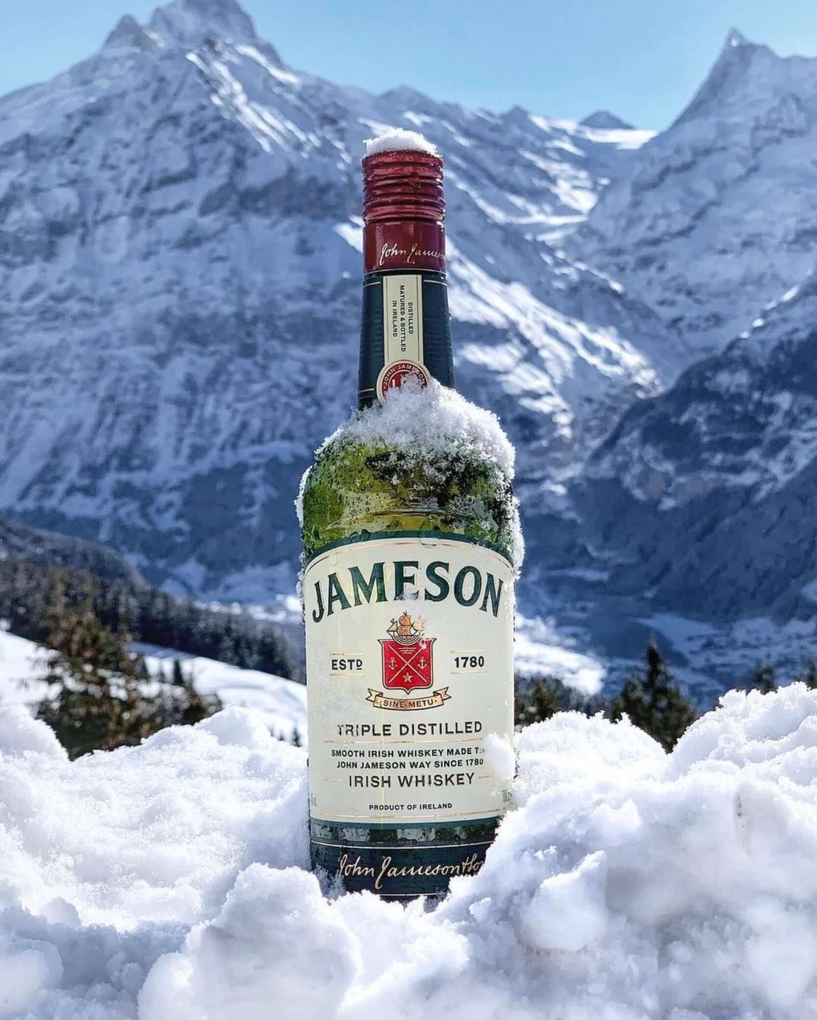 Jameson Irish Whiskey bottle in snow with snowy mountain backdrop, showcasing its iconic green bottle and smooth character.