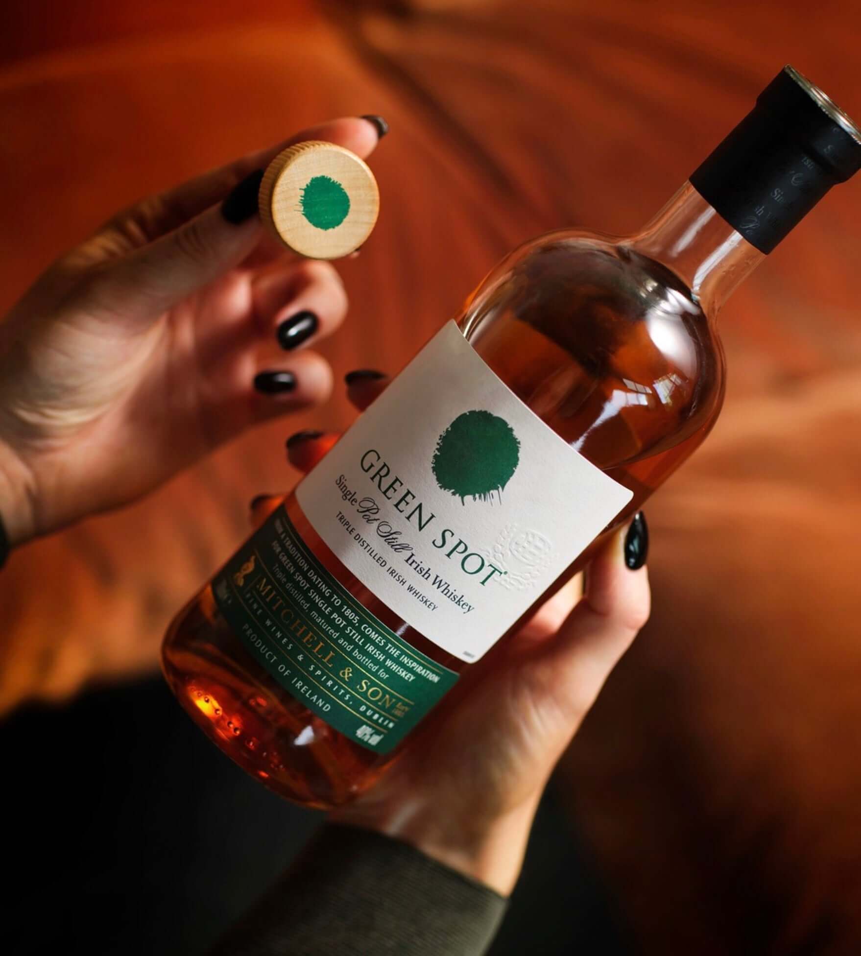 Green Spot Irish Whiskey bottle held by a person, featuring the brand logo and cork top on a wooden background.