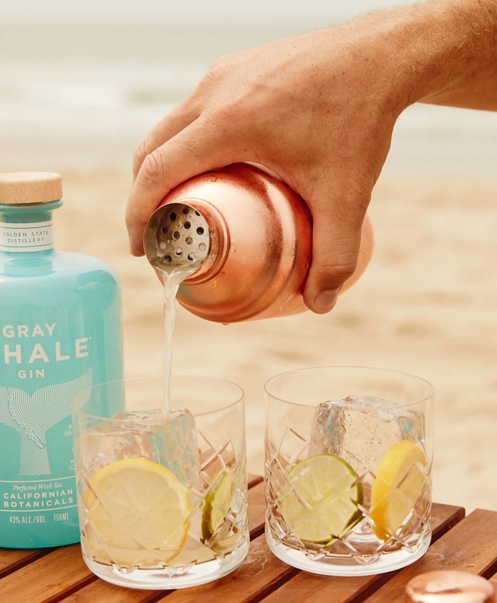 Hand pouring Gray Whale Gin into glasses with lime slices on a beach table.