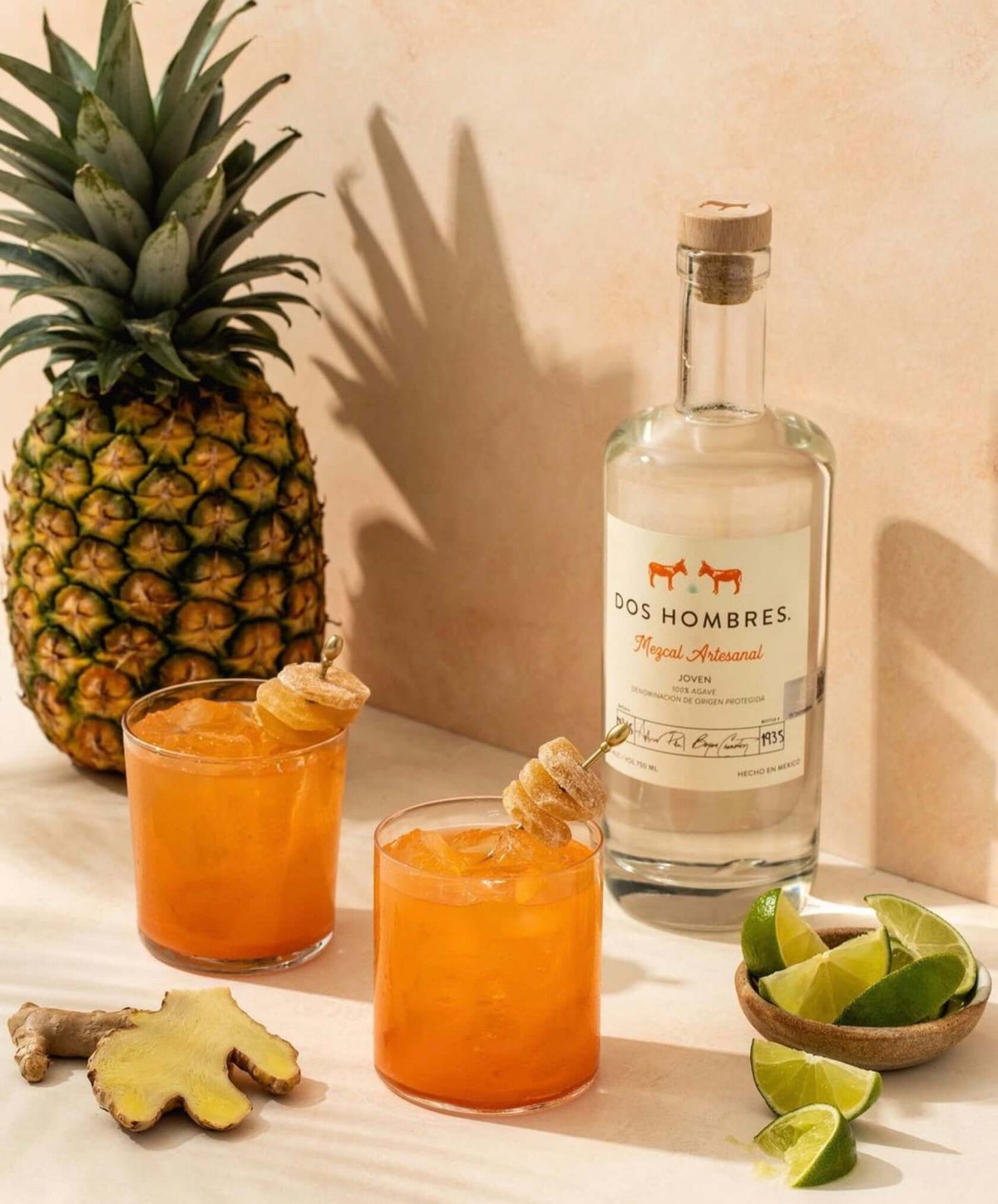 Dos Hombres Mezcal Espadin Joven with cocktails, pineapple, and lime, showcasing a vibrant tropical drink setup.