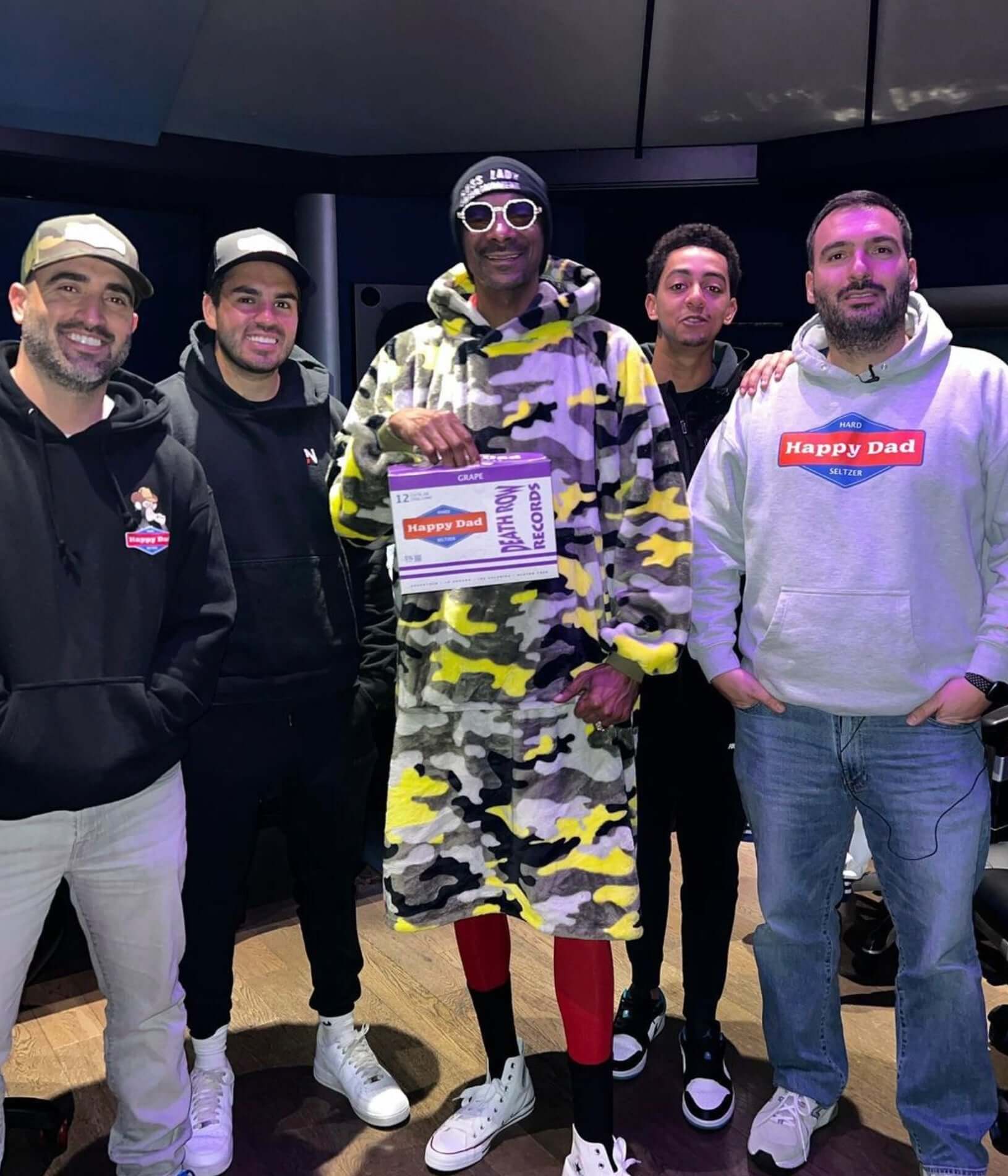 Group of five men posing together, one in a colorful camouflage outfit, promoting Happy Dad Grape Hard Seltzer.