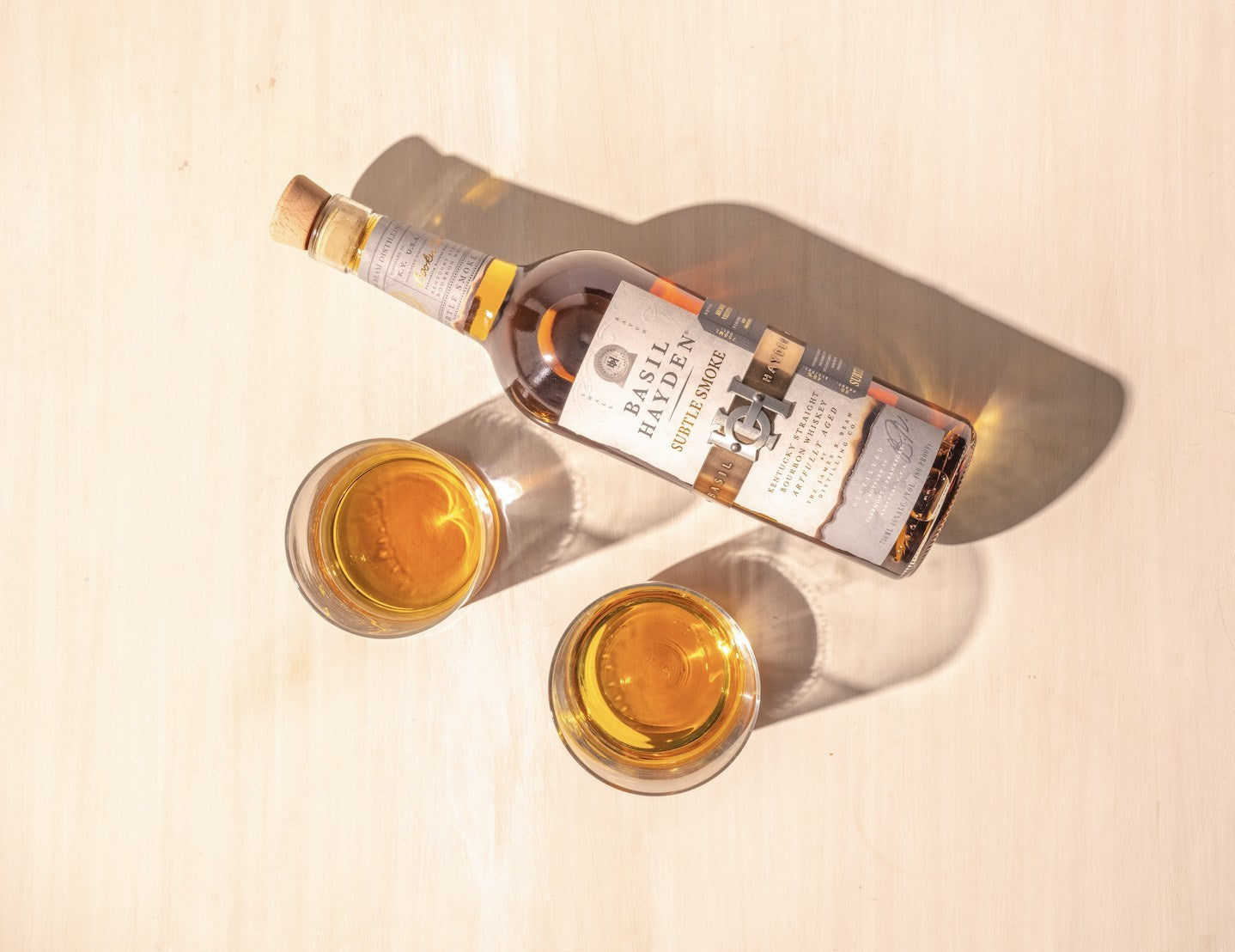 Basil Hayden Subtle Smoke whiskey bottle with two glasses of whiskey on a light surface.