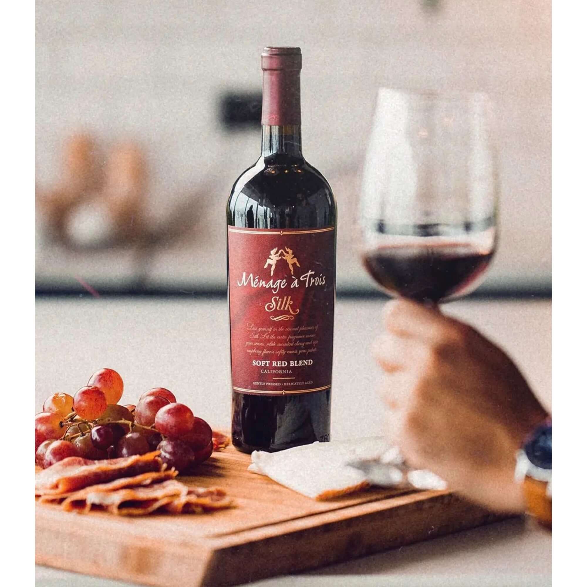 Ménage à Trois Silk Soft Red Wine bottle with glass, grapes, and charcuterie on a wooden board.