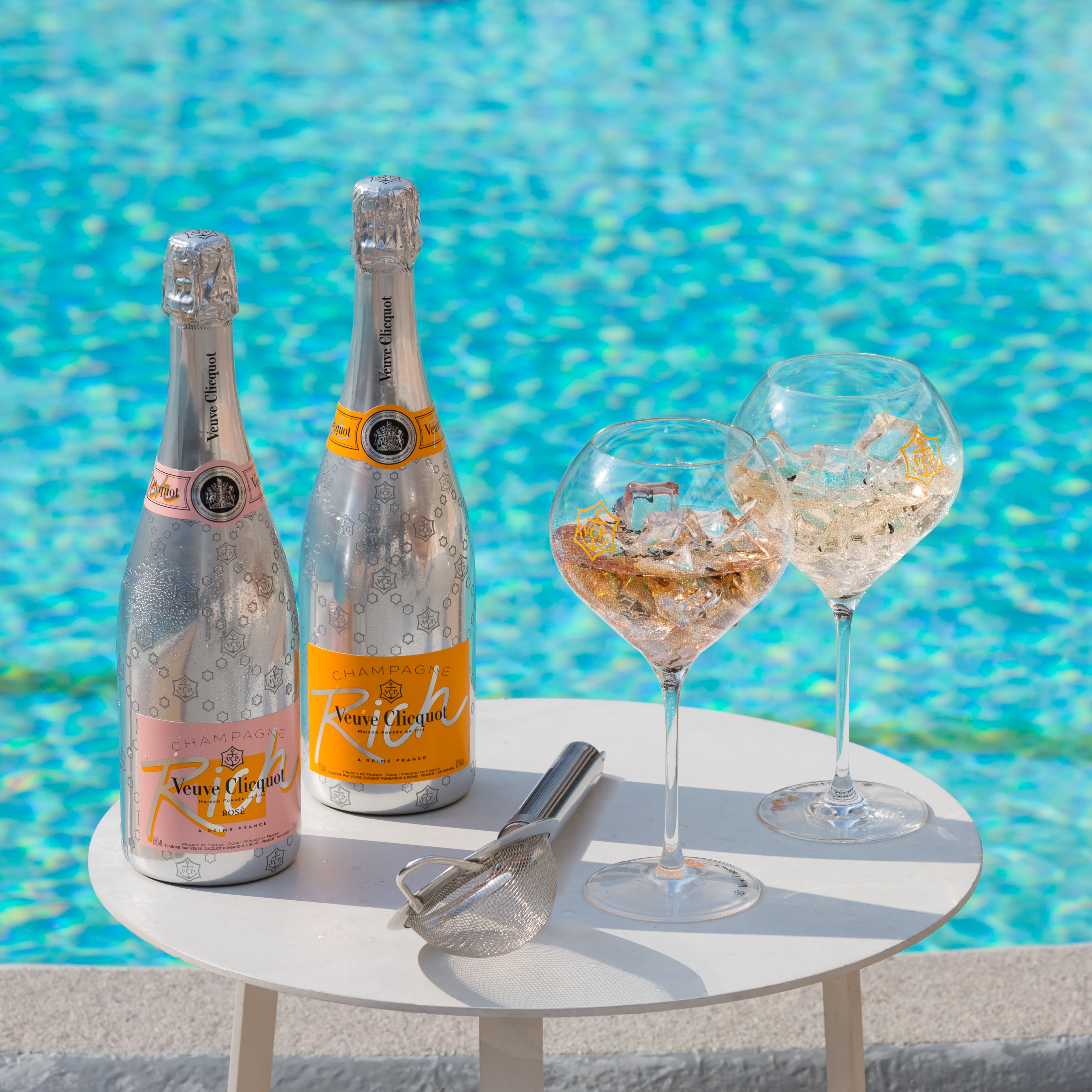 Two bottles of Veuve Clicquot Rich Rosé and Rich Champagne beside elegant glasses by a pool.
