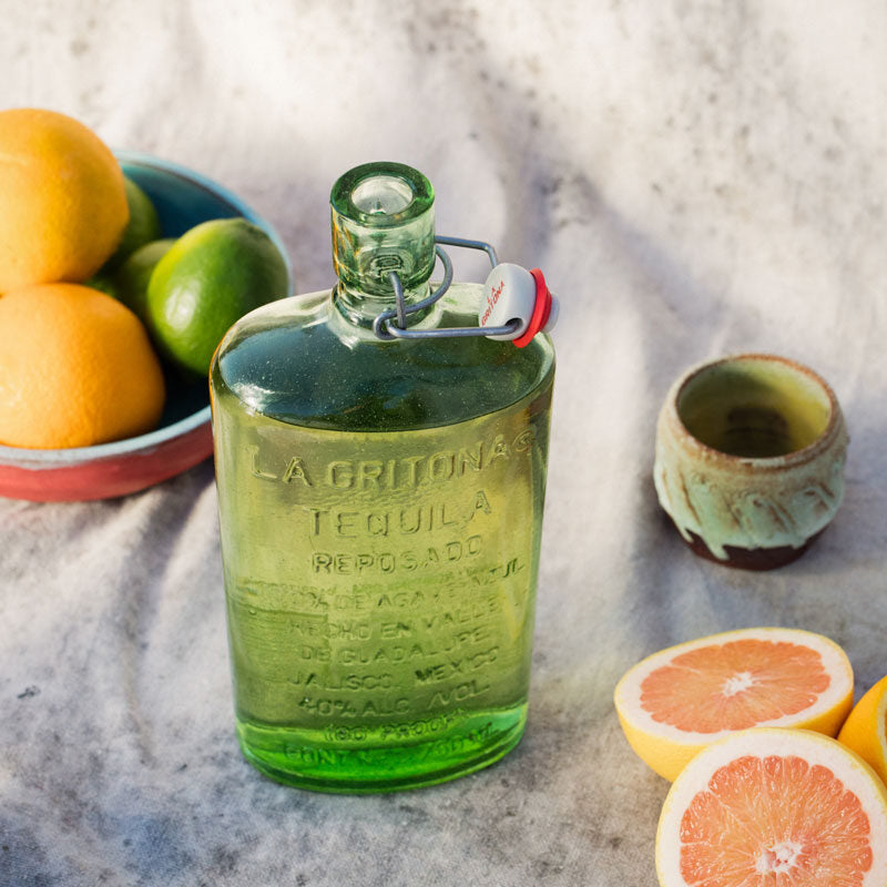 Bottle of La Gritona Reposado Tequila surrounded by fresh citrus fruits on a textured surface.