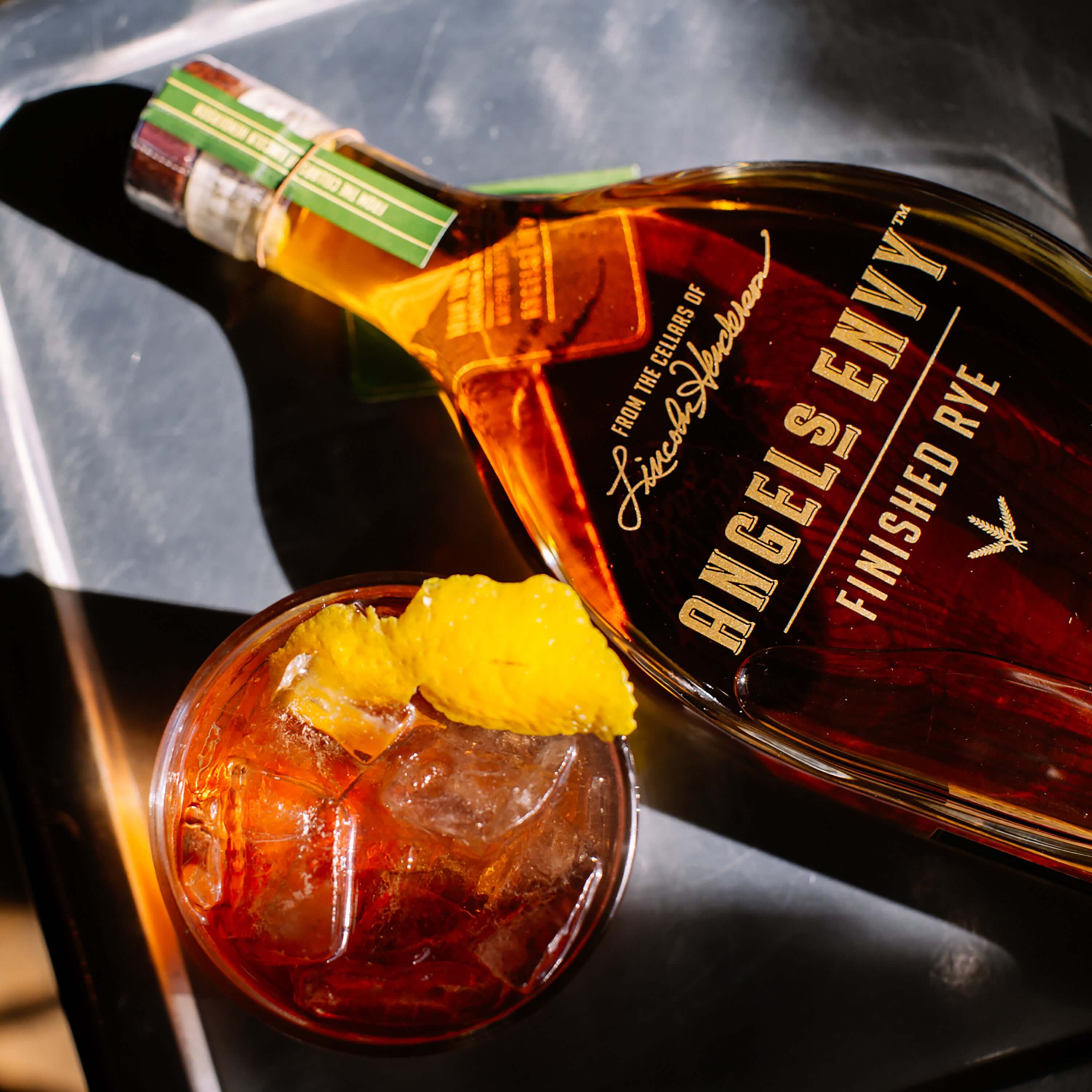 Angel's Envy Finished Rye Whiskey bottle next to a glass of cocktail with ice and citrus garnish.