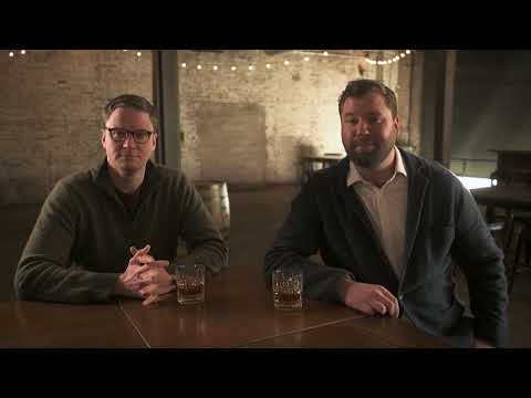 Two men sitting at a table with glasses of bourbon in a rustic distillery setting.