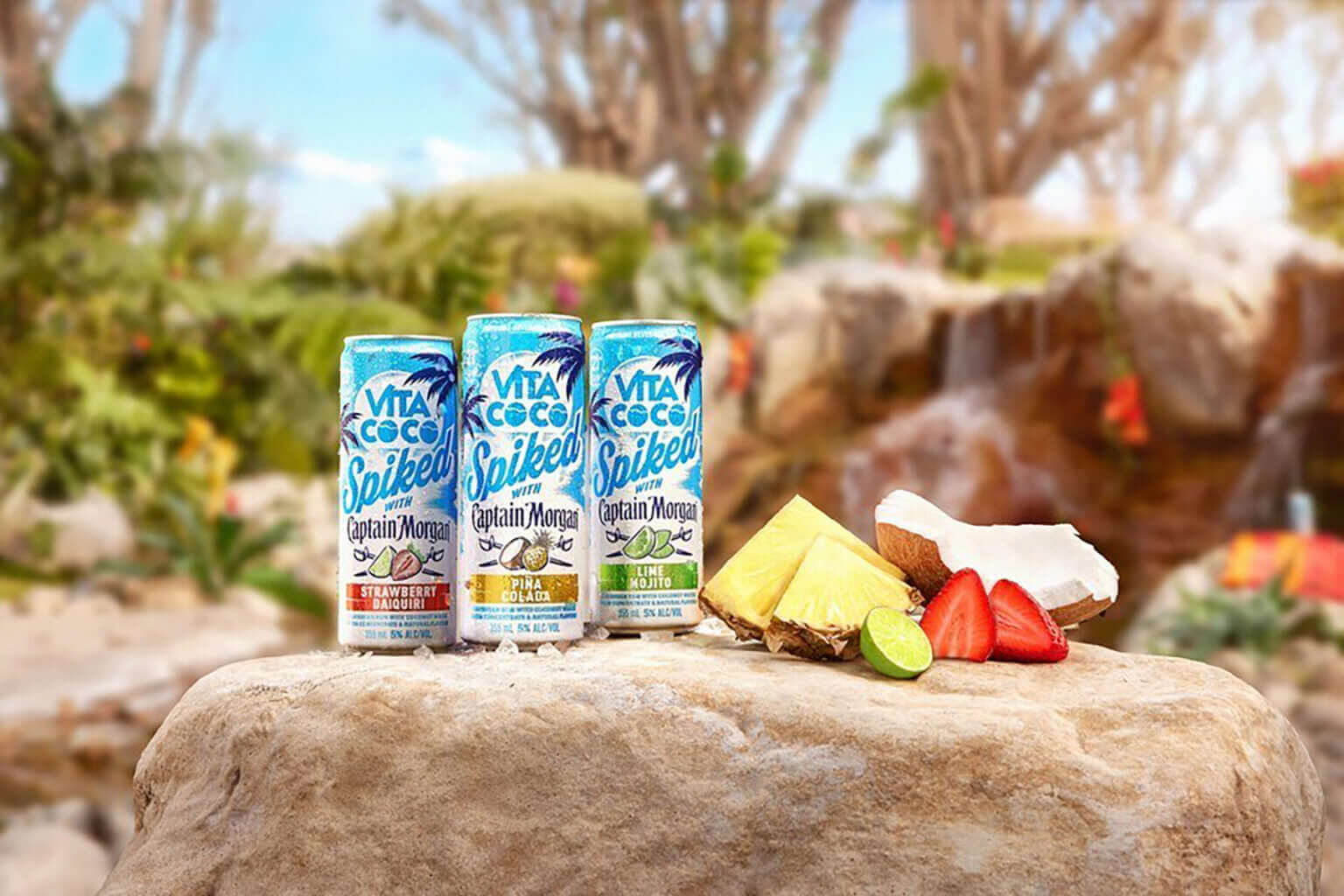 Vita Coco Spiked with Captain Morgan Piña Colada cans on a rock with tropical fruits in a vibrant outdoor setting.