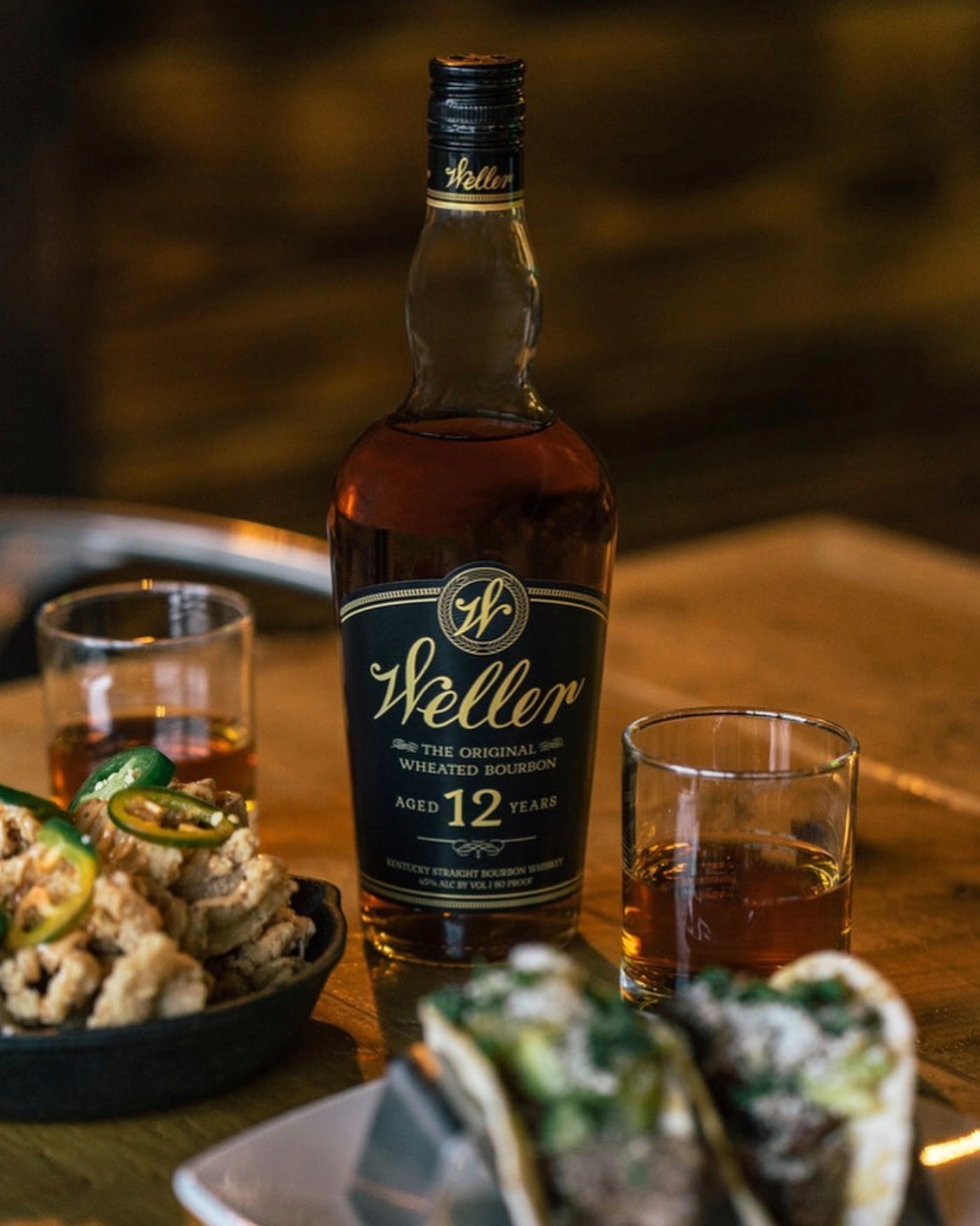 W.L. Weller 12 Year Wheated Bourbon bottle with drinks and food on a wooden table.