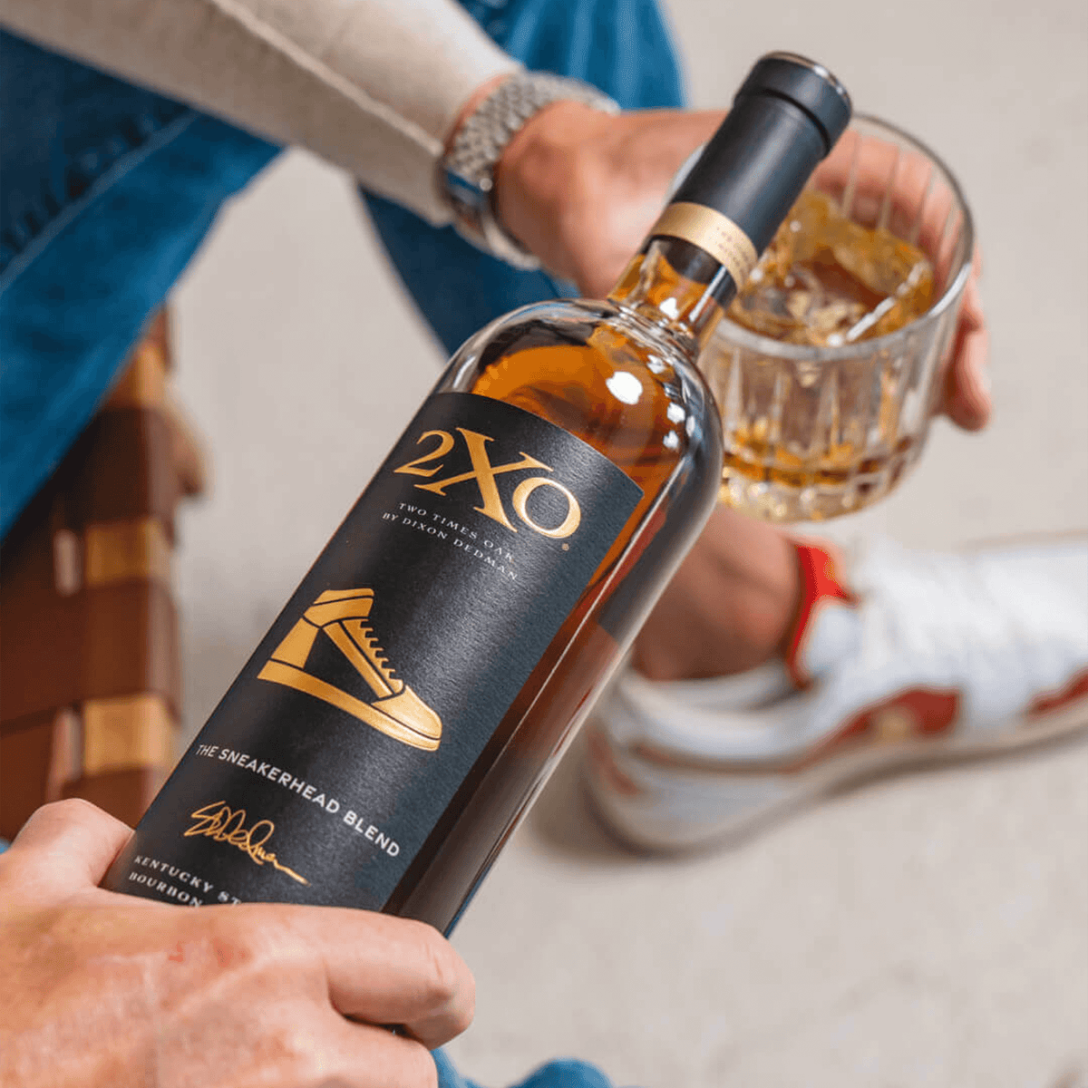 2XO The Sneakerhead Blend bourbon bottle beside a glass of bourbon, showcasing sneaker culture and style.