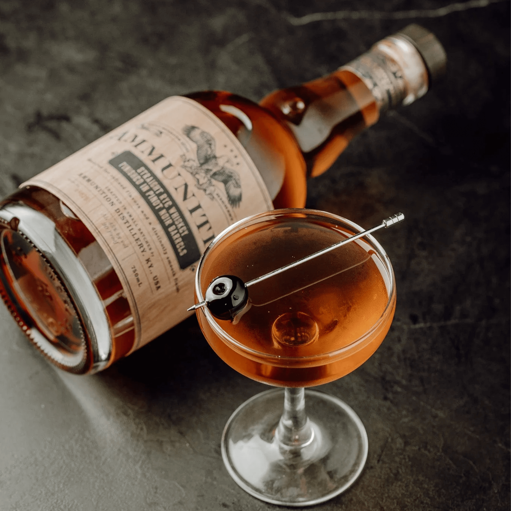 Ammunition Straight Rye Whiskey bottle beside a cocktail in a glass with an olive garnish on a dark countertop.