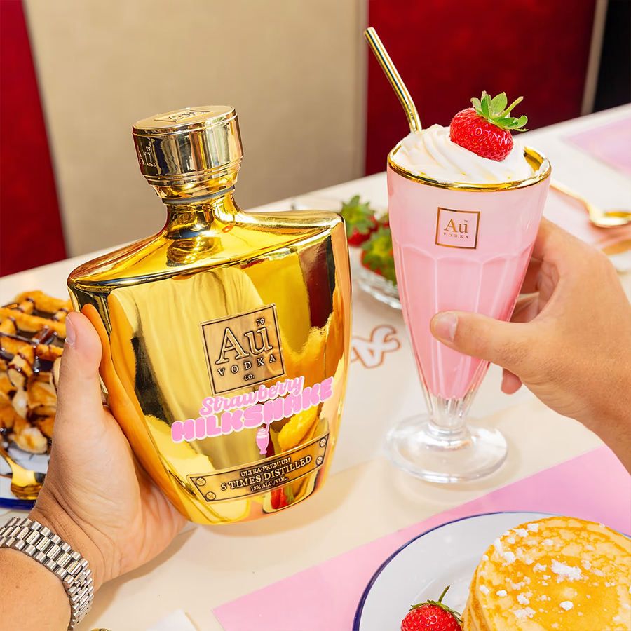 Person holding Au Vodka Strawberry Milkshake Liqueur 700ml bottle next to a strawberry milkshake dessert.