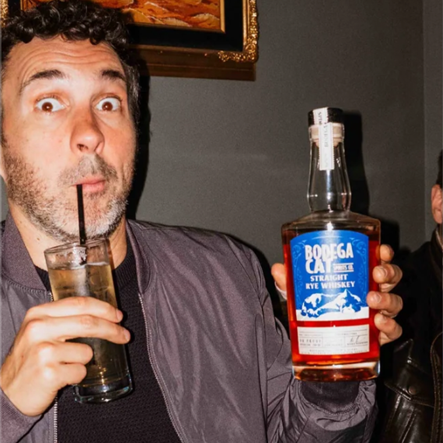 Person holding a bottle of Bodega Cat Straight Rye Whiskey with a drink in hand, smiling in a lively setting.