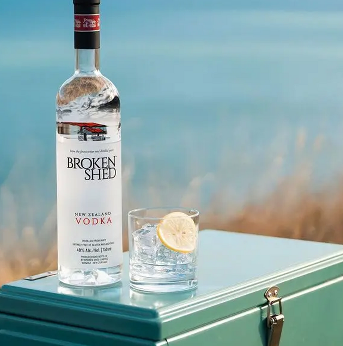 Bottle of Broken Shed Vodka next to a glass of ice and lemon on a blue cooler by a scenic backdrop.