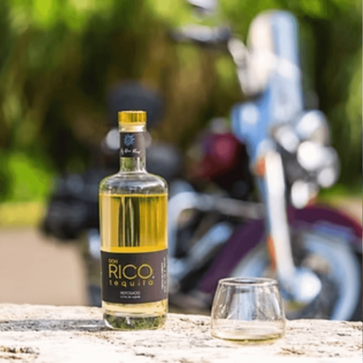 Bottle of Don Rico Reposado Tequila with a glass on a stone surface, motorcycle in the background.