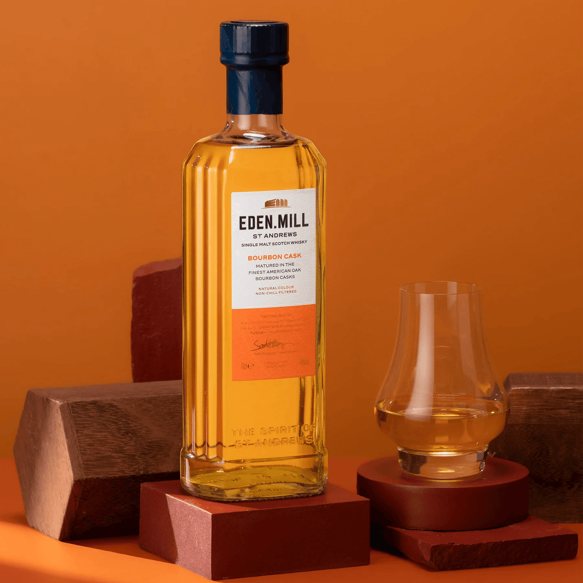 Eden Mill Bourbon Cask Single Malt Scotch 700ml with tasting glass on wooden surfaces against an orange backdrop.