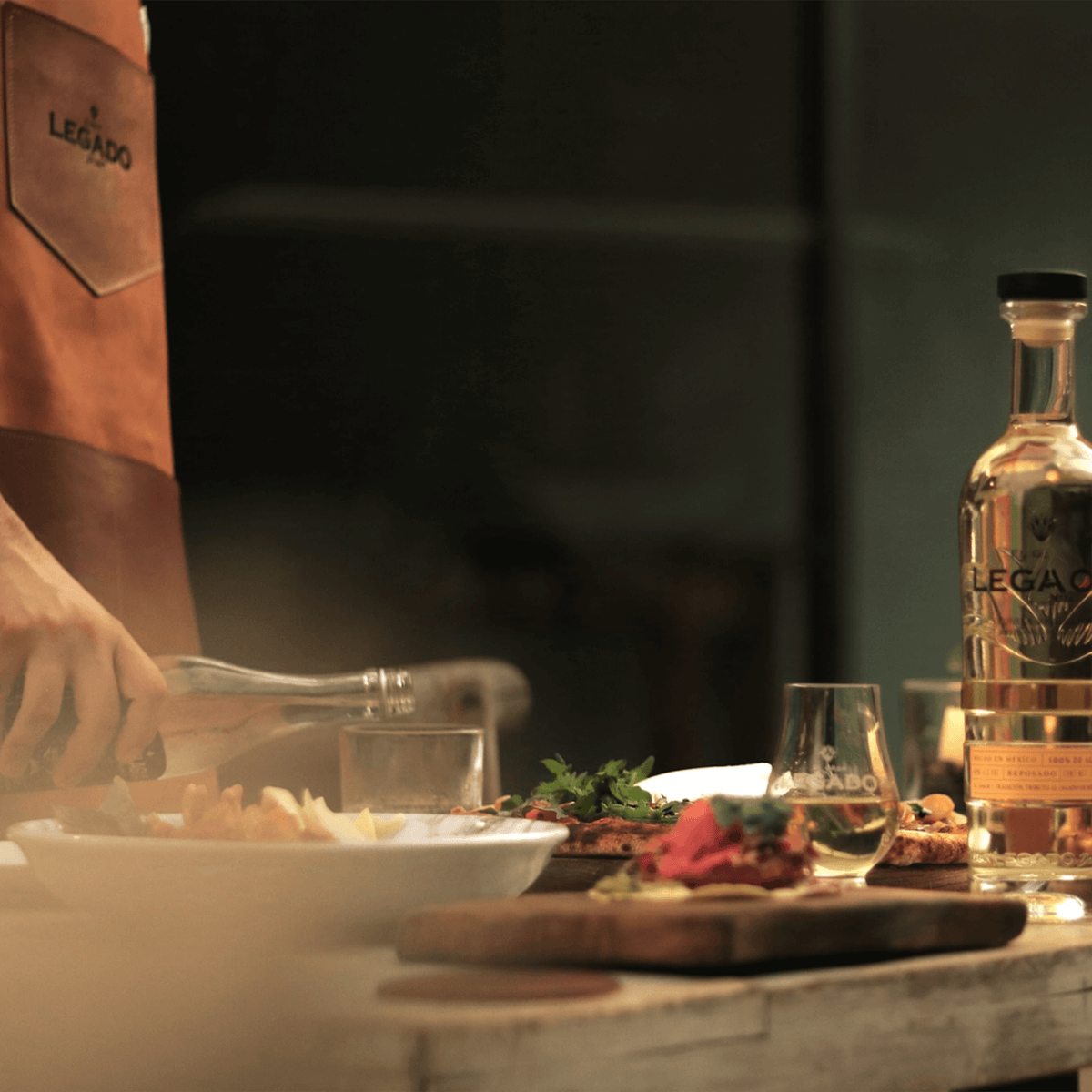 Glass of El Gran Legado Añejo Tequila on a wooden table with gourmet dishes, showcasing a culinary experience.