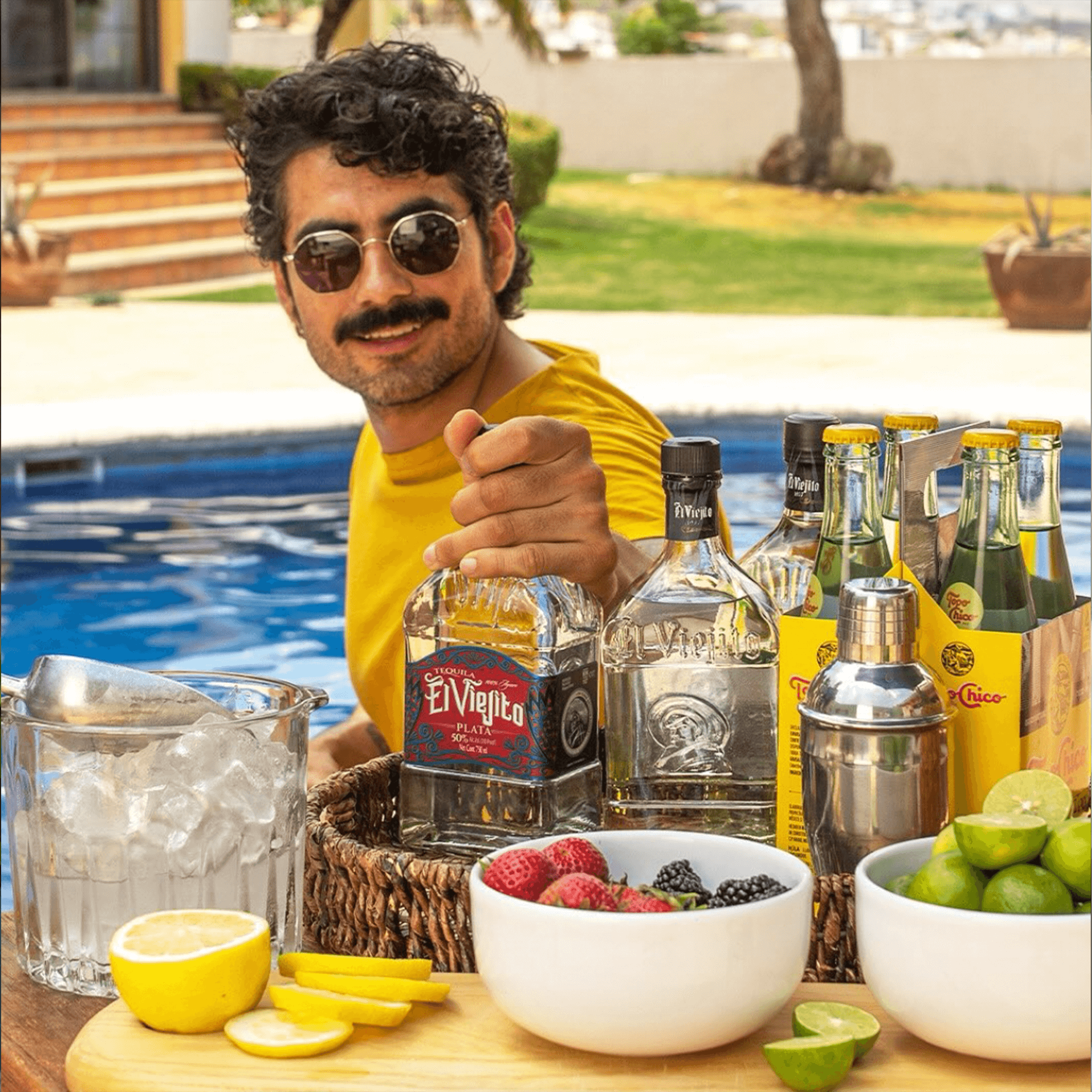 Man enjoying El Viejito Plata 50% tequila outdoors by a pool, surrounded by ingredients for cocktails.