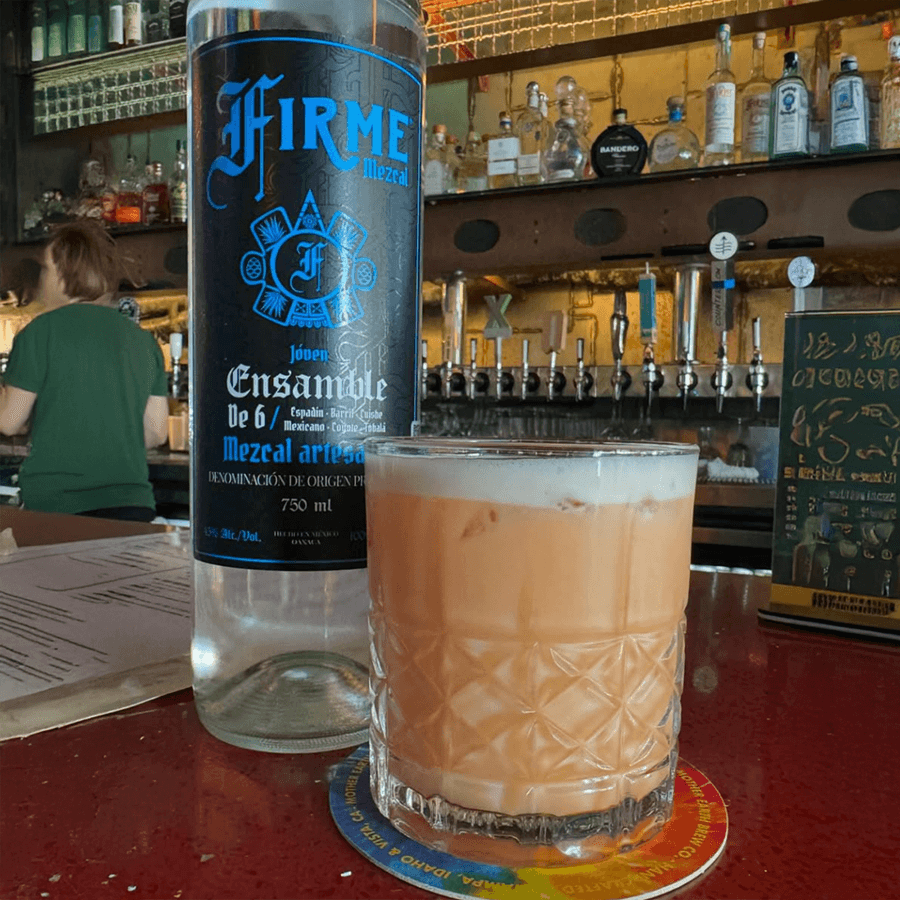 Firme Mezcal Joven Ensamble de 6 bottle next to a cocktail in a vibrant bar setting.