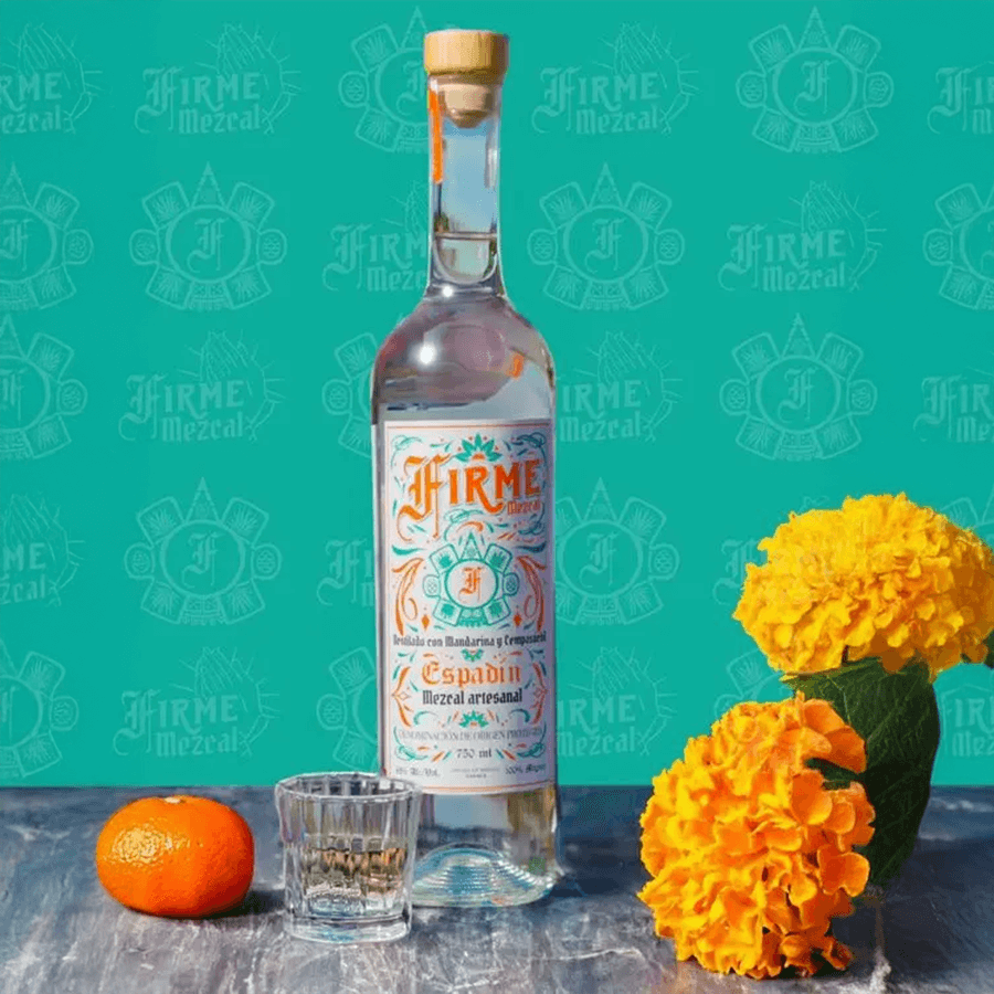 Firme Mezcal Joven Espadín bottle with cempasúchil flowers and tangerine on a wooden table.