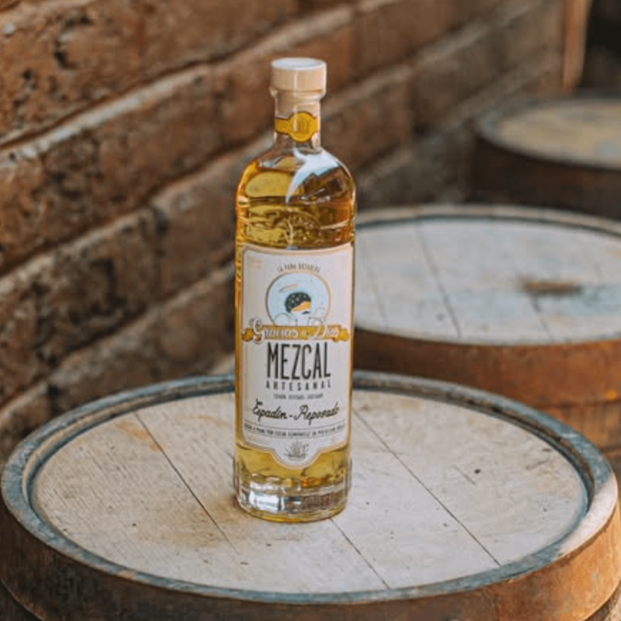 Gracias A Dios Espadin Reposado Mezcal bottle on wooden barrels, showcasing its gold color and artisanal label.