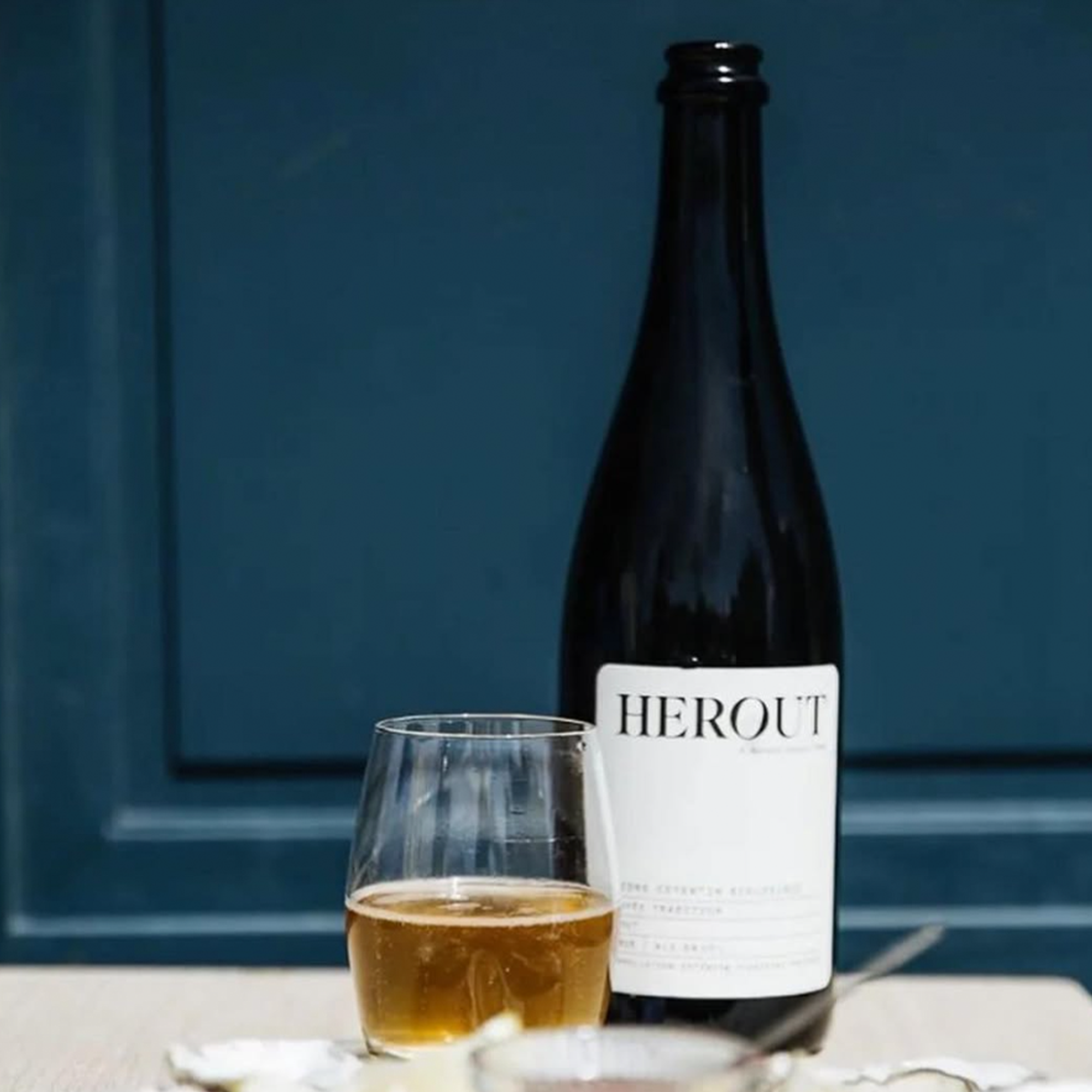 Bottle of Herout Grande Cuvée Extra Brut 2020 with a glass of cider on a table, showcasing its elegant appearance.