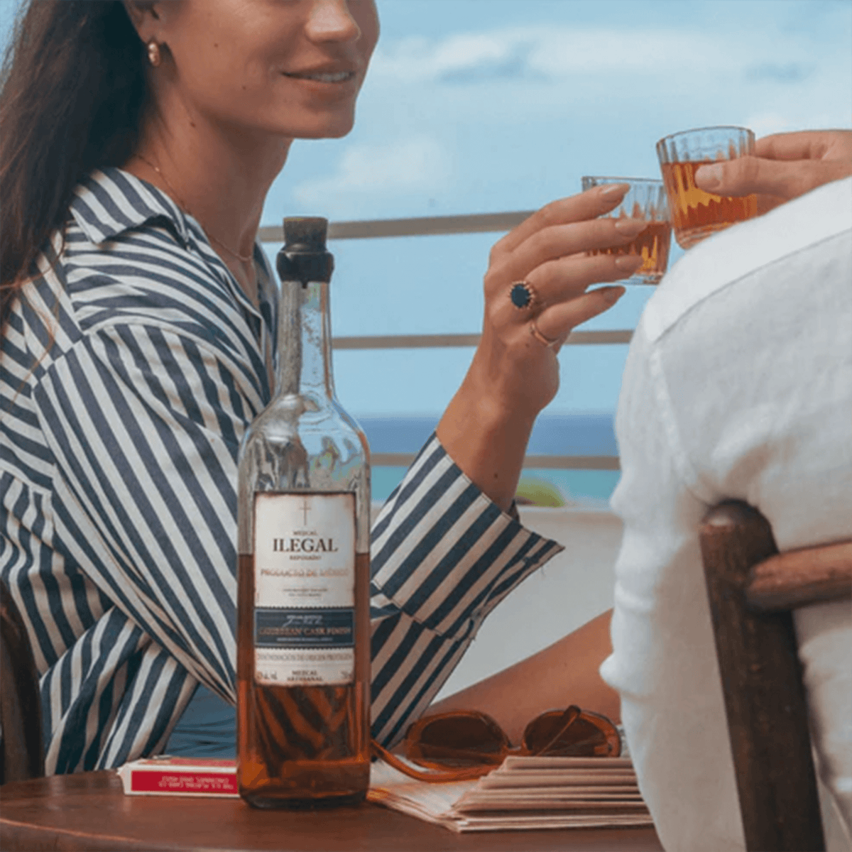 Friends toasting with glasses of Ilegal Reposado Caribbean Cask Finish Mezcal by the ocean.