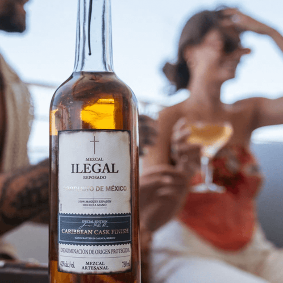 Ilegal Reposado Caribbean Cask Finish Mezcal bottle with two people enjoying cocktails on a sunny day.