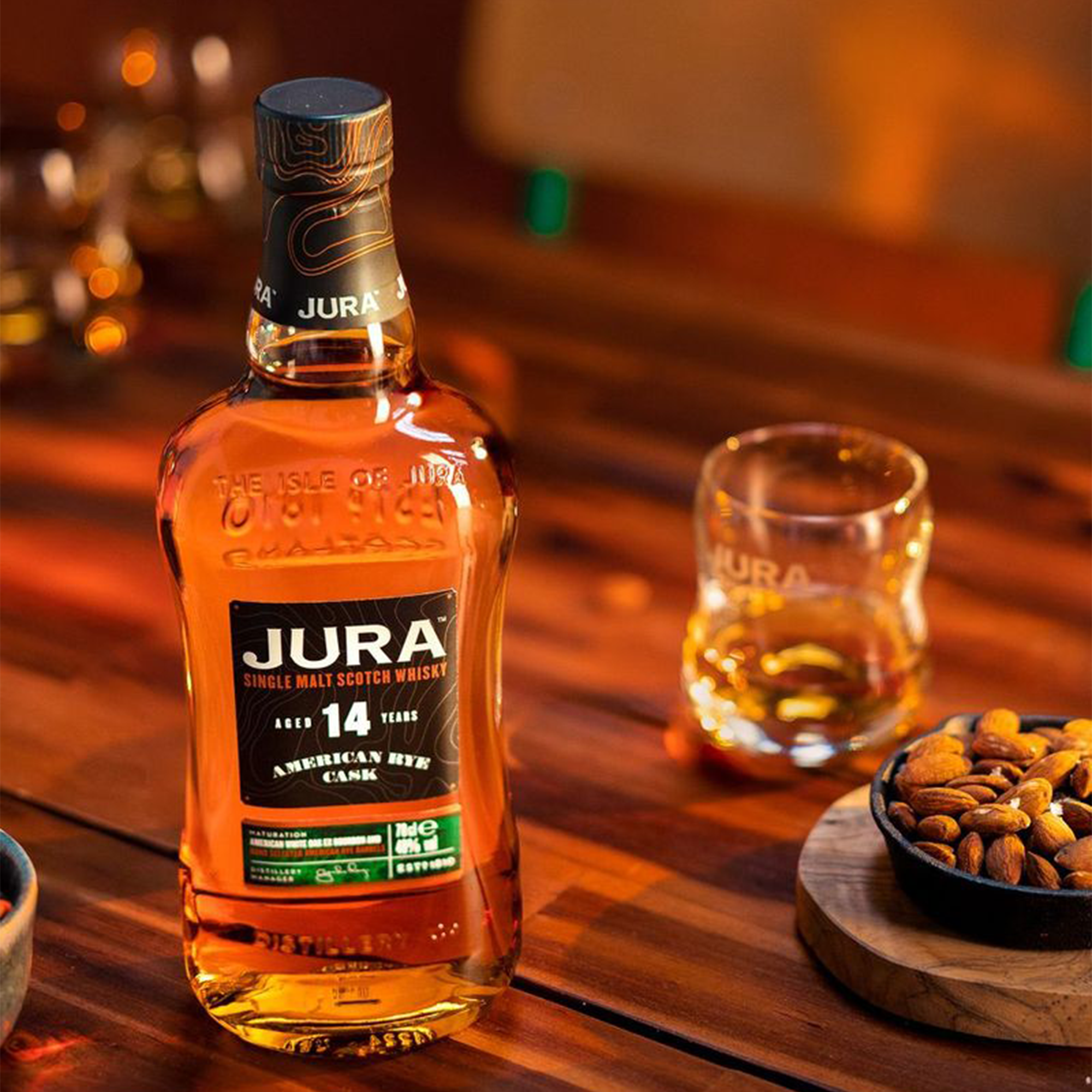 Bottle of Jura 14 Year American Rye Cask whiskey with a glass and almonds on a wooden table.