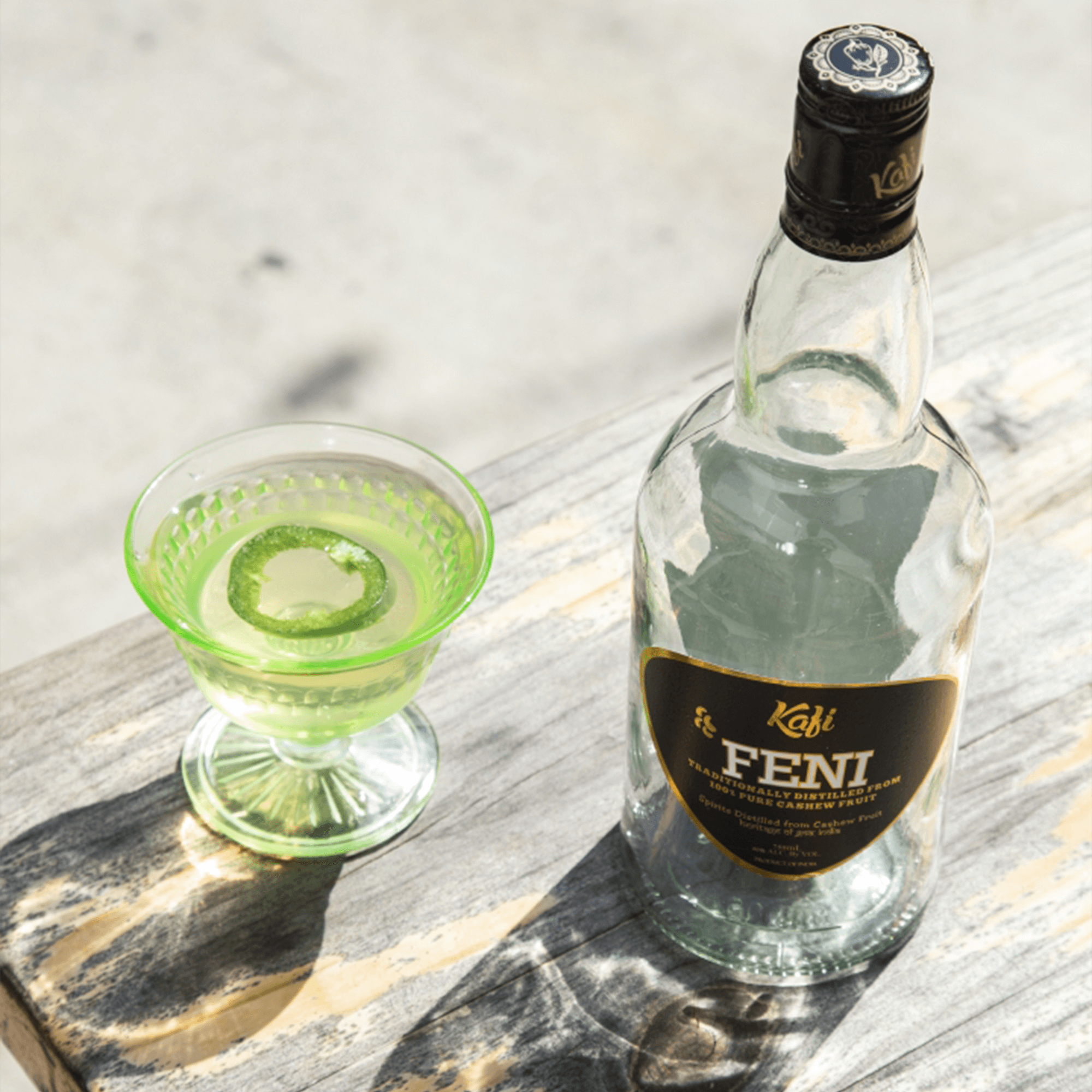 Kafi Cashew Feni brandy bottle next to a vibrant green cocktail in a decorative glass.