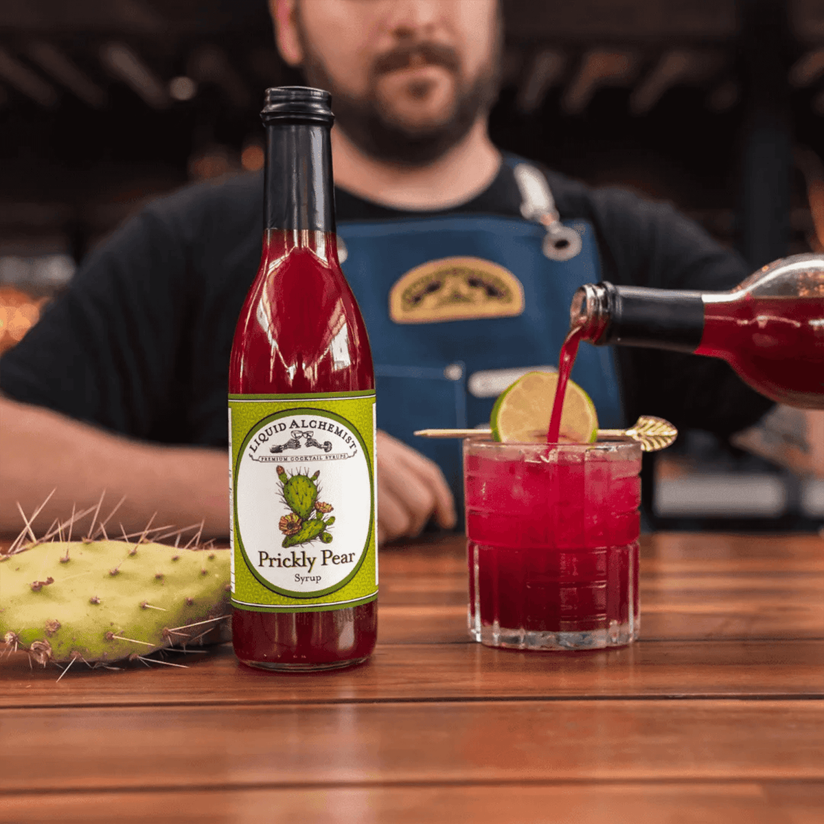 Liquid Alchemist Prickly Pear Syrup bottle beside a vibrant prickly pear cocktail garnished with lime.