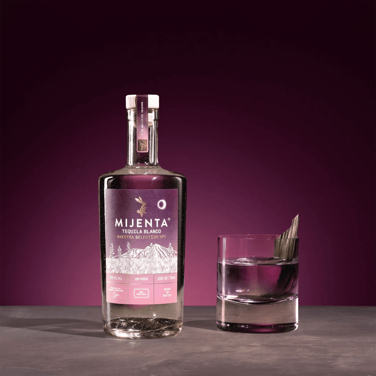 Mijenta Maestra Selection No 1 Blanco Tequila bottle next to a glass with an agave leaf garnish on a purple background.