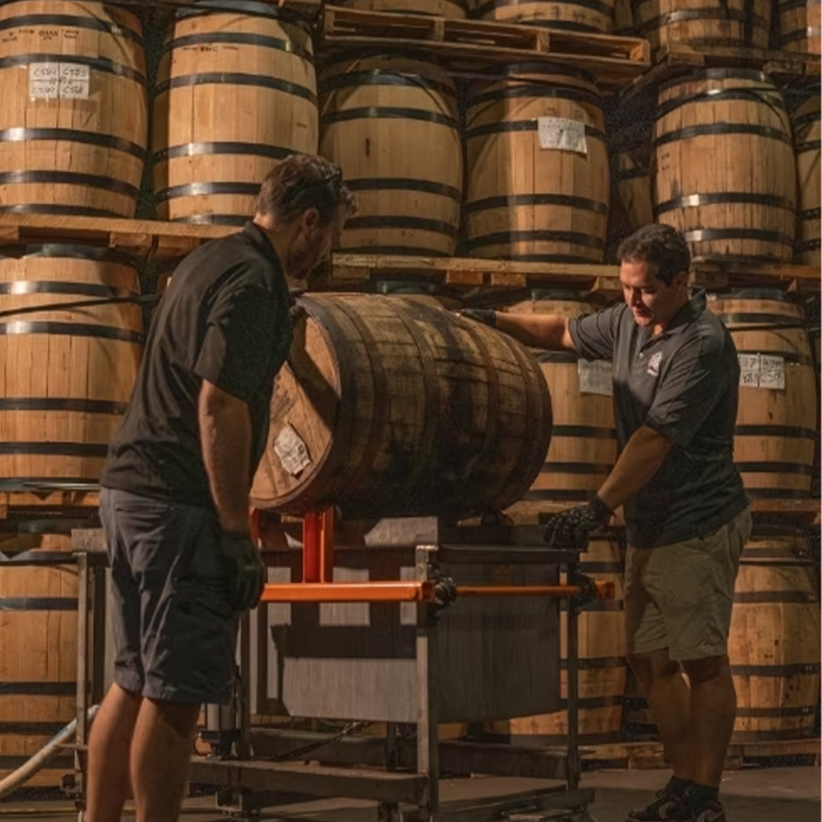 Two distillers inspecting a bourbon barrel in a warehouse filled with stacked barrels for blending.